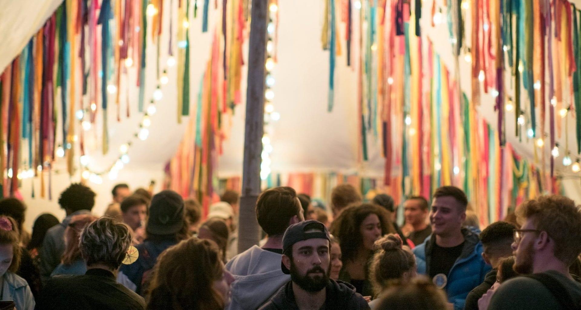 Crowd of young people at an outdoor event, gathering under a pole with lines of lights and streamers hanging from it.