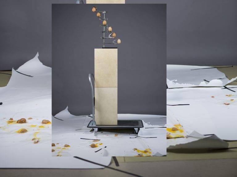 A photo of a piece of artwork. Ripped sheets of paper lie on the floor. A luggage trolley is in the middle with a pair of blocks stacked on top. On top of these blocks is a series of clamps, each holding an egg.