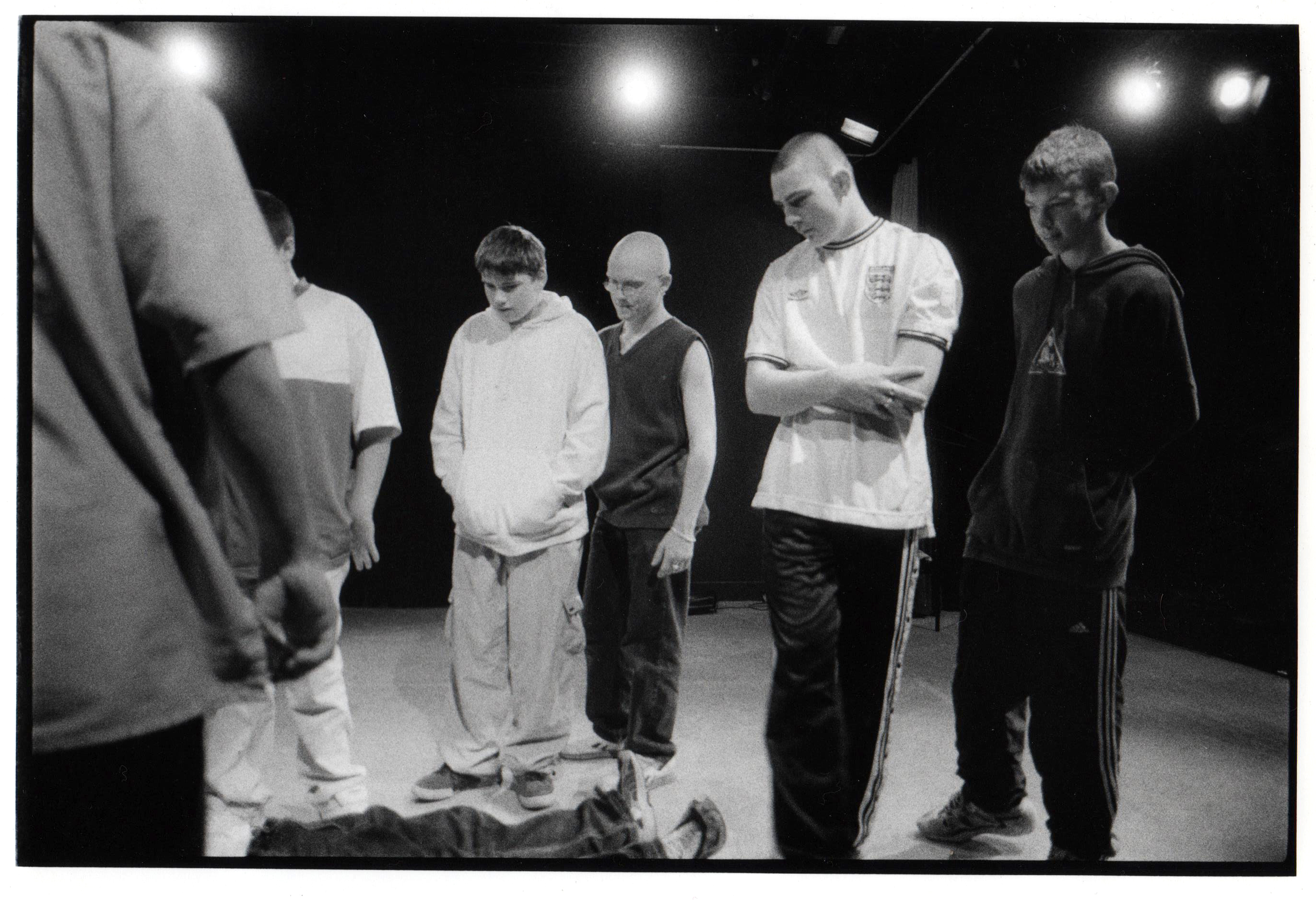 A group of young men on stage rehearsing a play, standing and looking at one of their classmates lying on the stage.