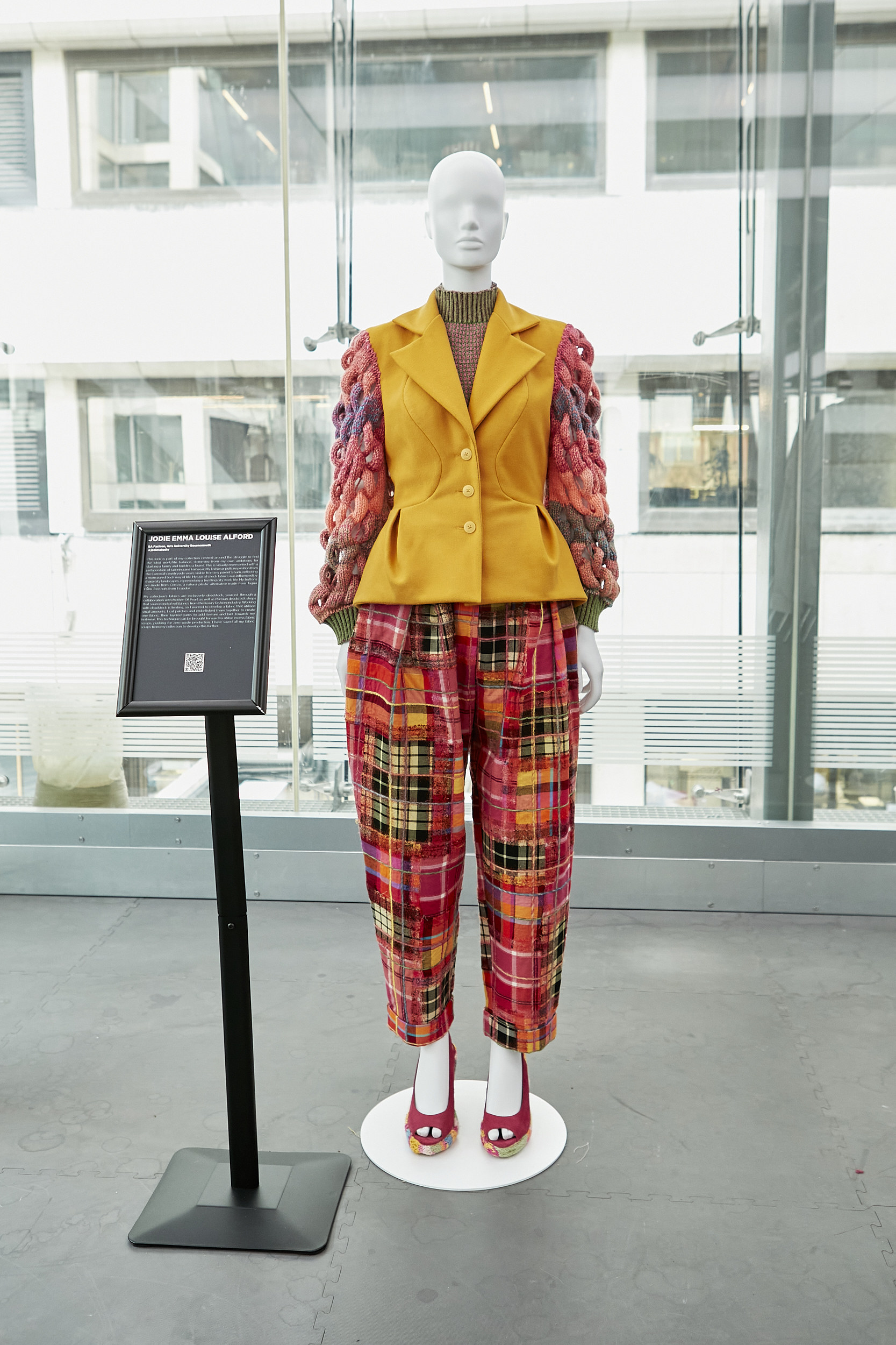 Mannequin wearing a yellow coat with red fabric-woven arms and a mixed pattern tartan skirt.