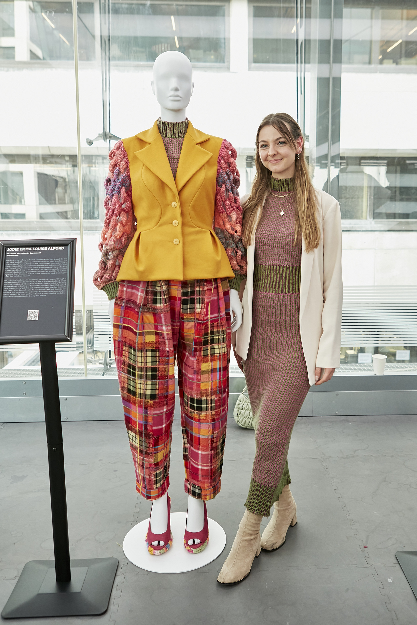 AUB student J Alford stood next to a mannequin wearing a yellow coat with red fabric-woven arms and a mixed pattern tartan skirt.