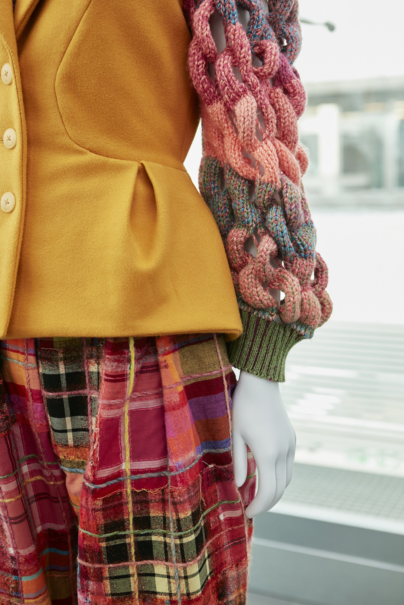 Yellow coat with red fabric-woven arms and a mixed pattern tartan skirt on a mannequin.