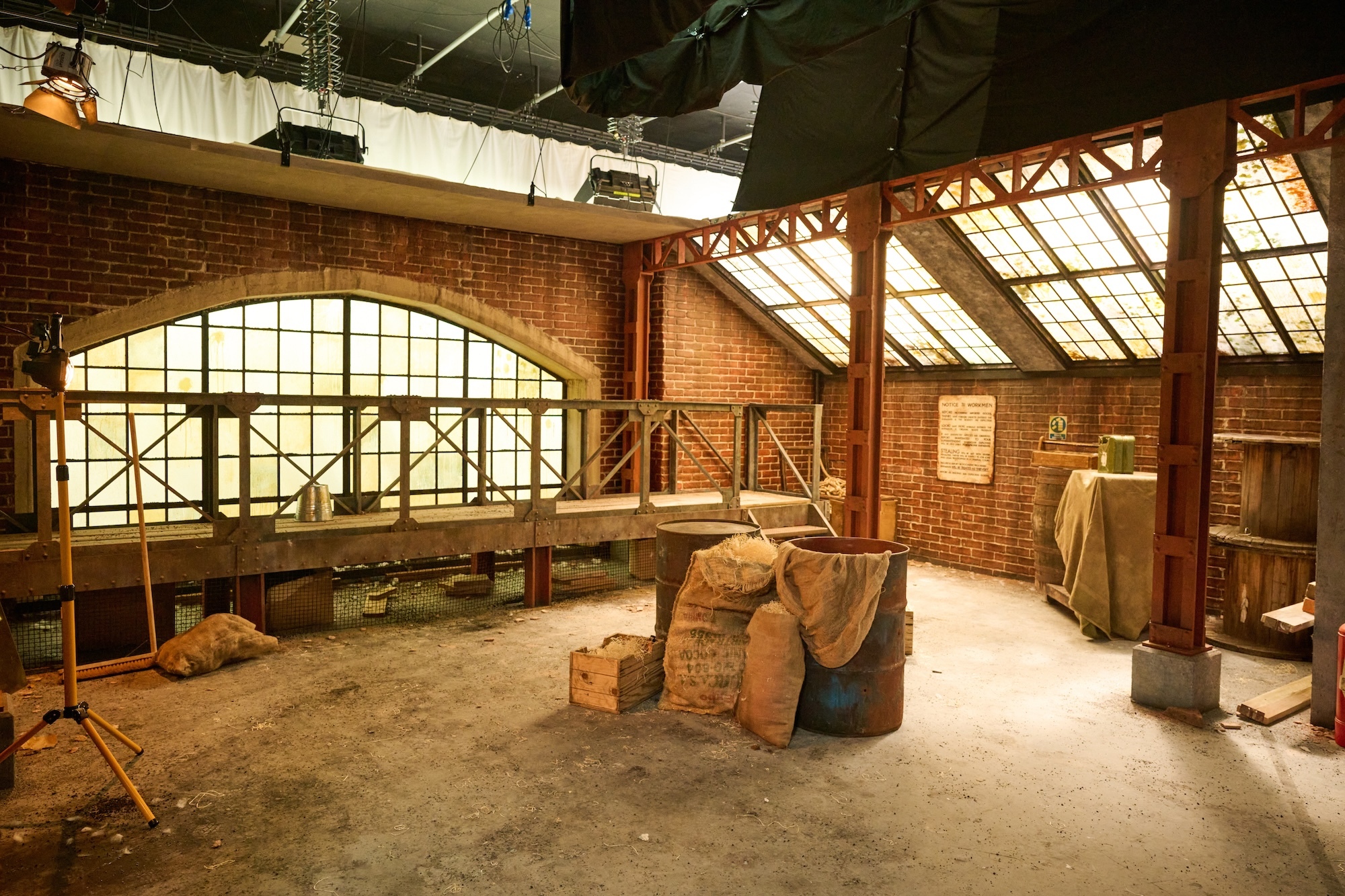 Film set designed as an industrial brick building interior with exposed timber roof structure, metal railings on upper level, and large sacks on concrete floor.