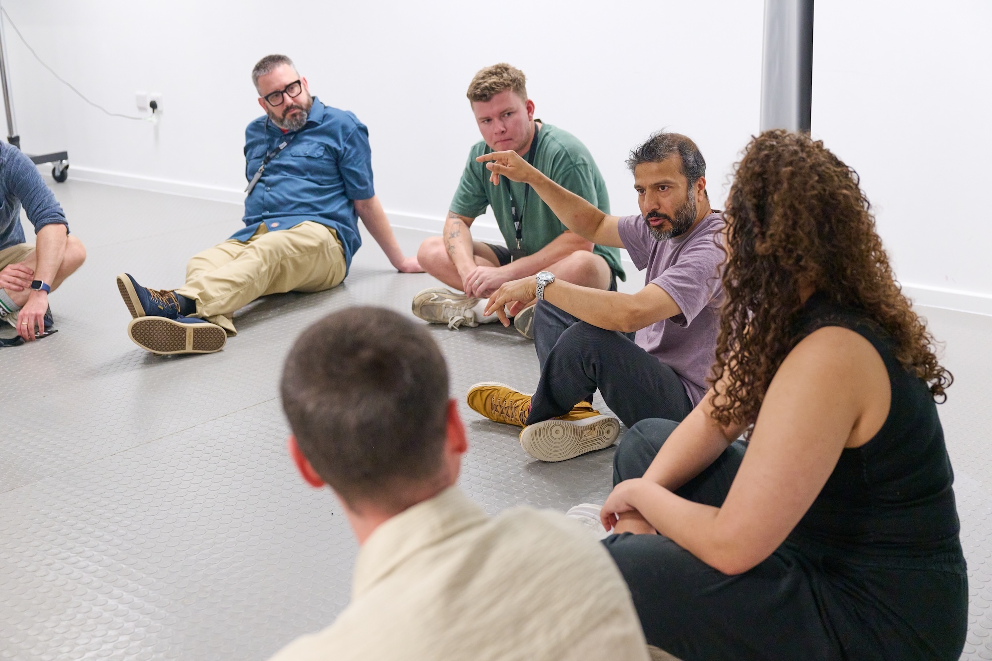 Six people sitting in circle on white floor during group discussion or meeting in bright room with white walls.