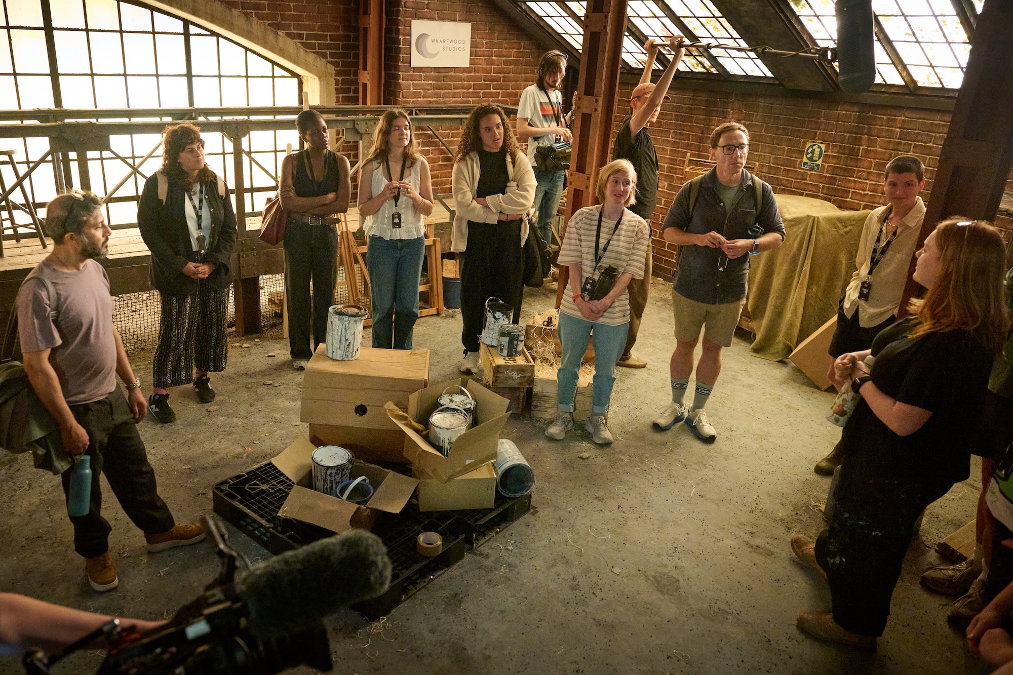 Group of people gathered on a film set designed like a brick warehouse space with large windows, film equipment on floor, industrial setting with concrete floors.