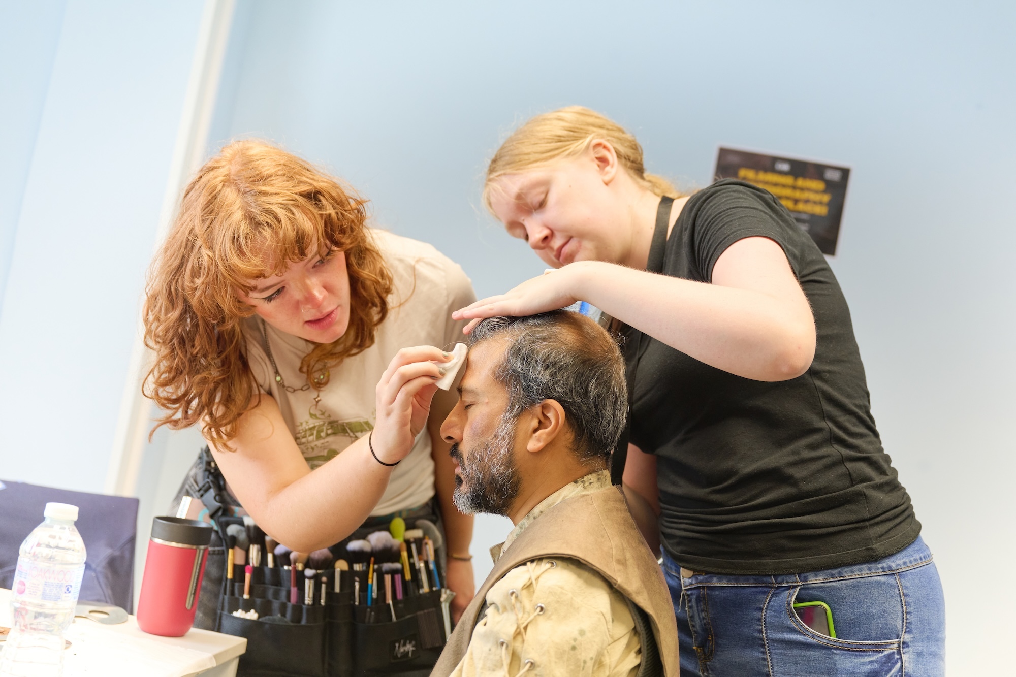 Two make-up artists applying prosthetics to seated man's face and head, with supplies on table beside them.