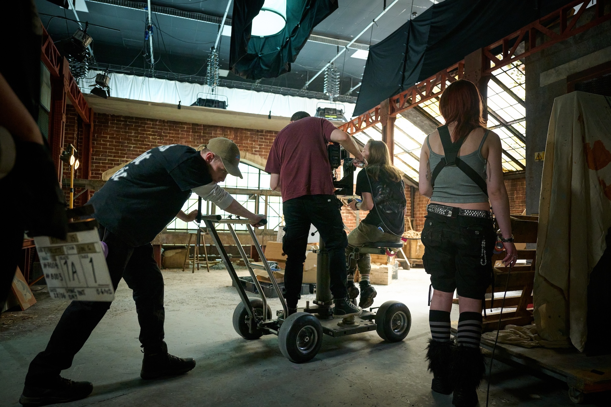Film crew operating camera dolly in warehouse with brick walls, exposed ceiling, and industrial lighting equipment.