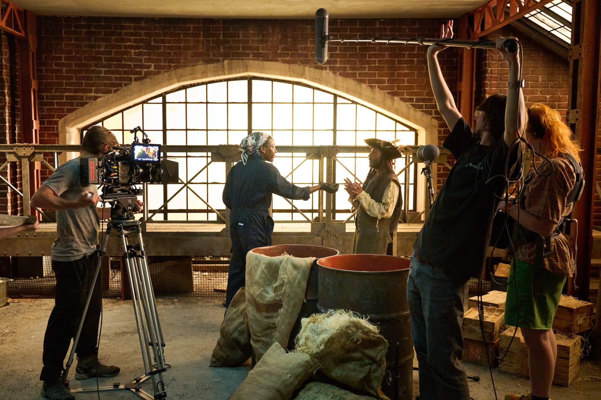 Film crew with cameras and equipment in brick warehouse with large arched window, bags and barrels on floor.