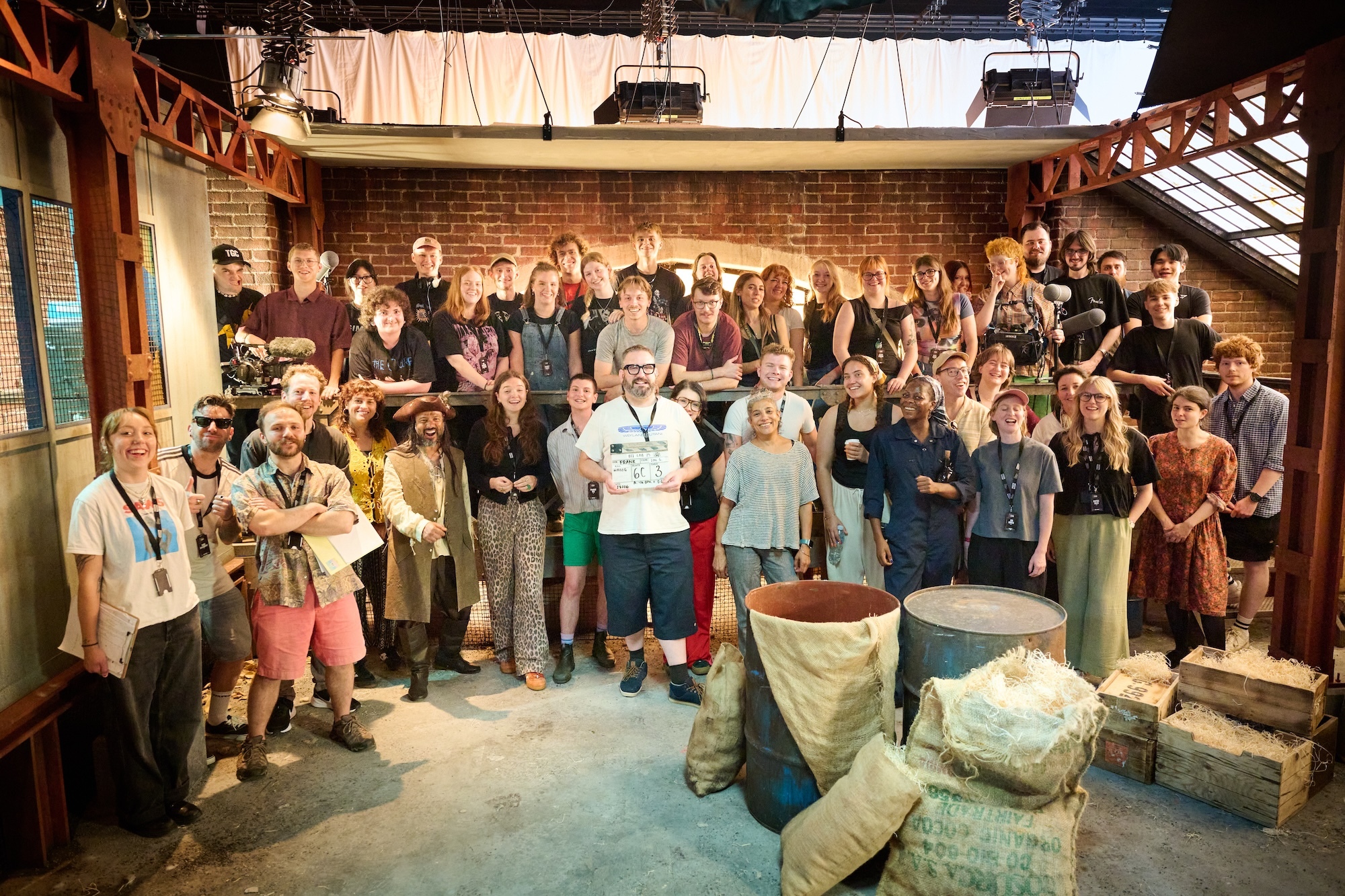 Large group of people gathered in industrial-style venue with exposed brick walls, steel beams, and covered equipment or furniture.
