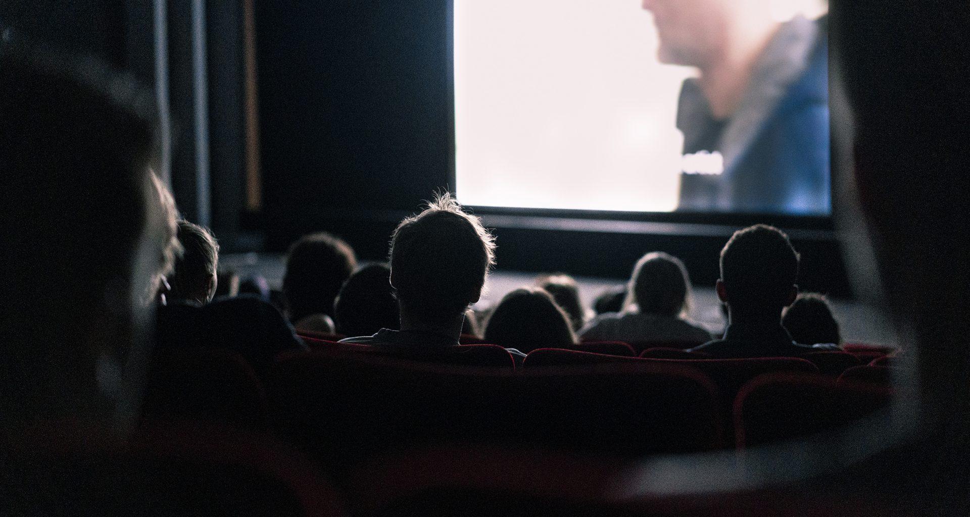 The backs of heads sitting in a movie theatre in darkness, with a film in progress on the screen in front of them