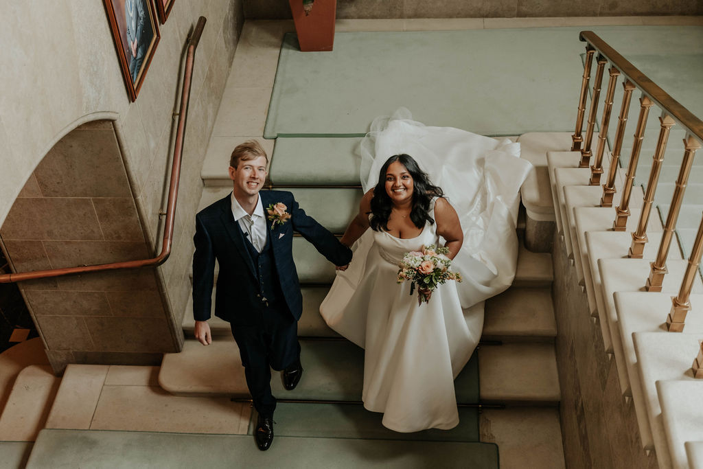Bride in white gown and groom in dark suit sitting on marble staircase with wooden banister in elegant interior setting.