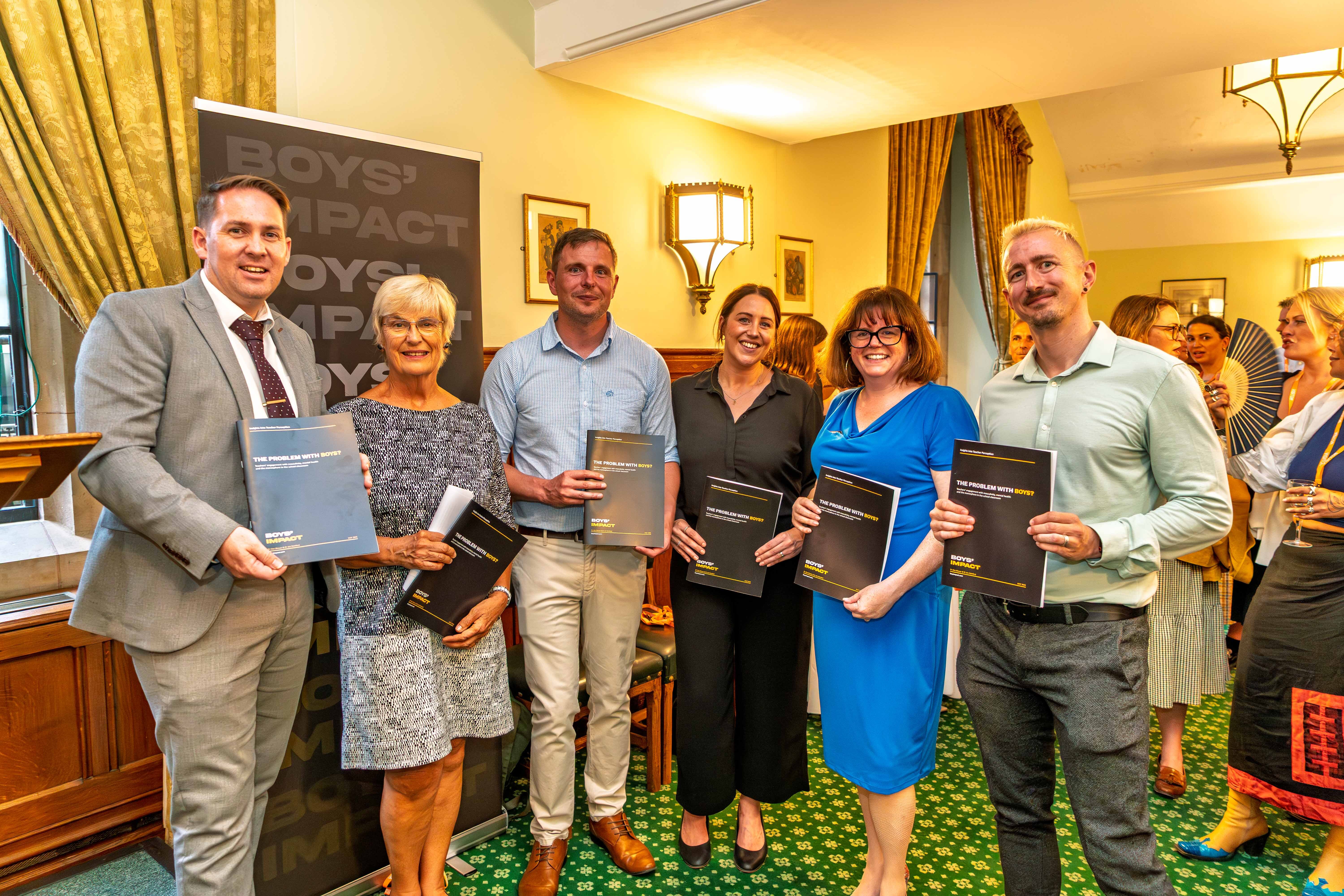 Six people holding packages for Boys' Impact in a formal room at the House of Commons with green carpet, yellow walls, and golden curtains.