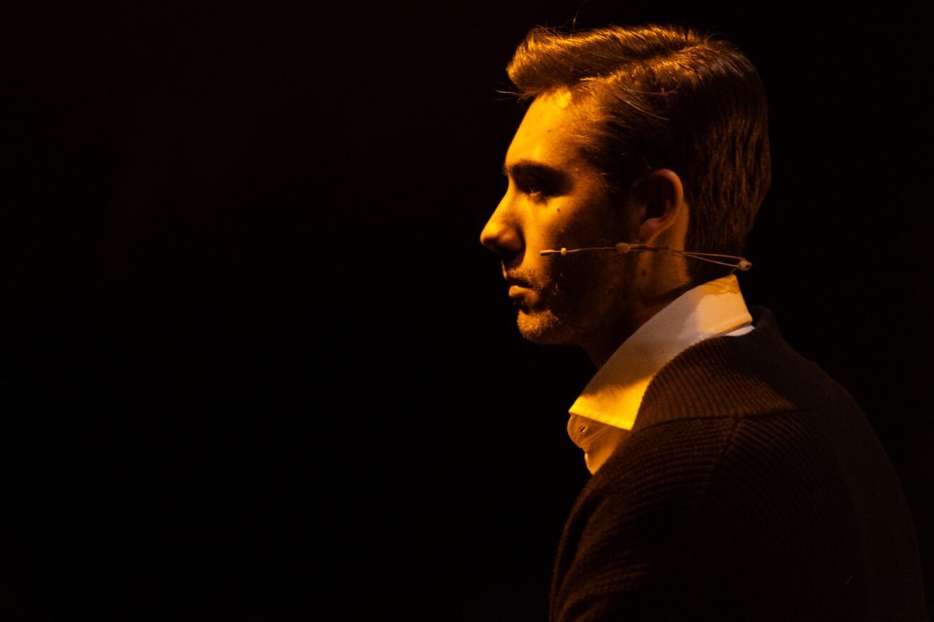 An actor under orange stage lights on a dark background.