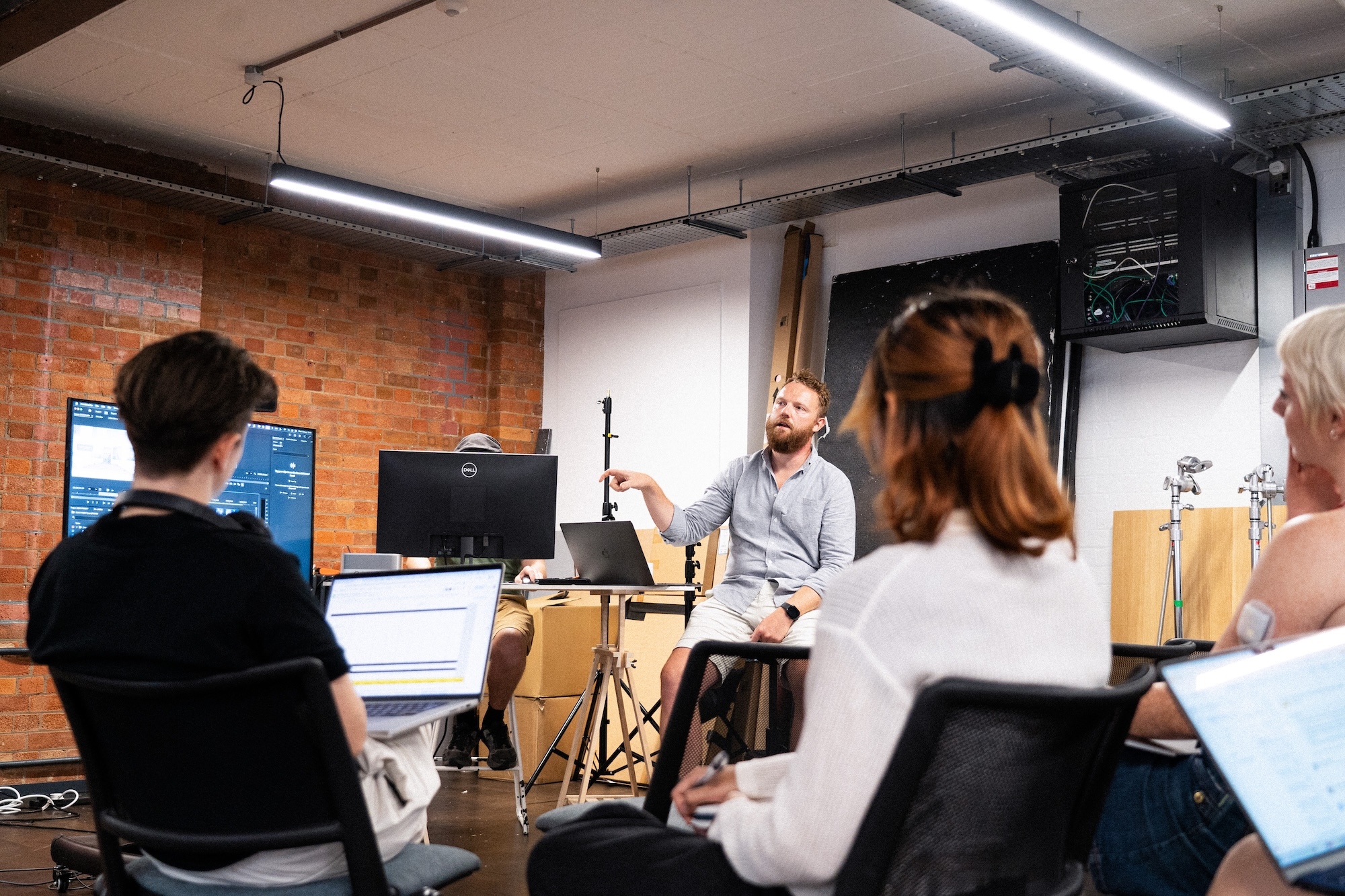 Three students viewed seated from behind while listening to a lecturer explaining something in a studio space.