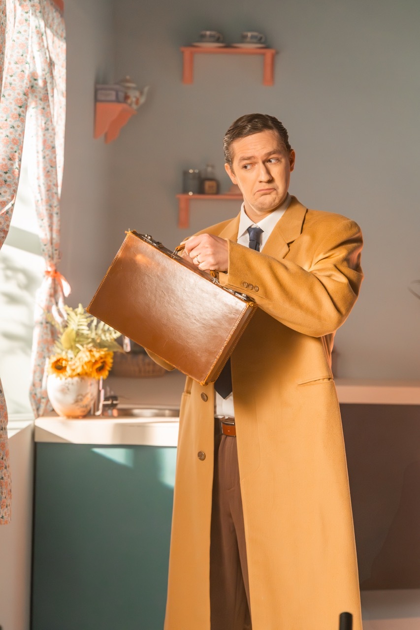 Man in a yellow coat holding a briefcase in a room with floral curtains and shelves.