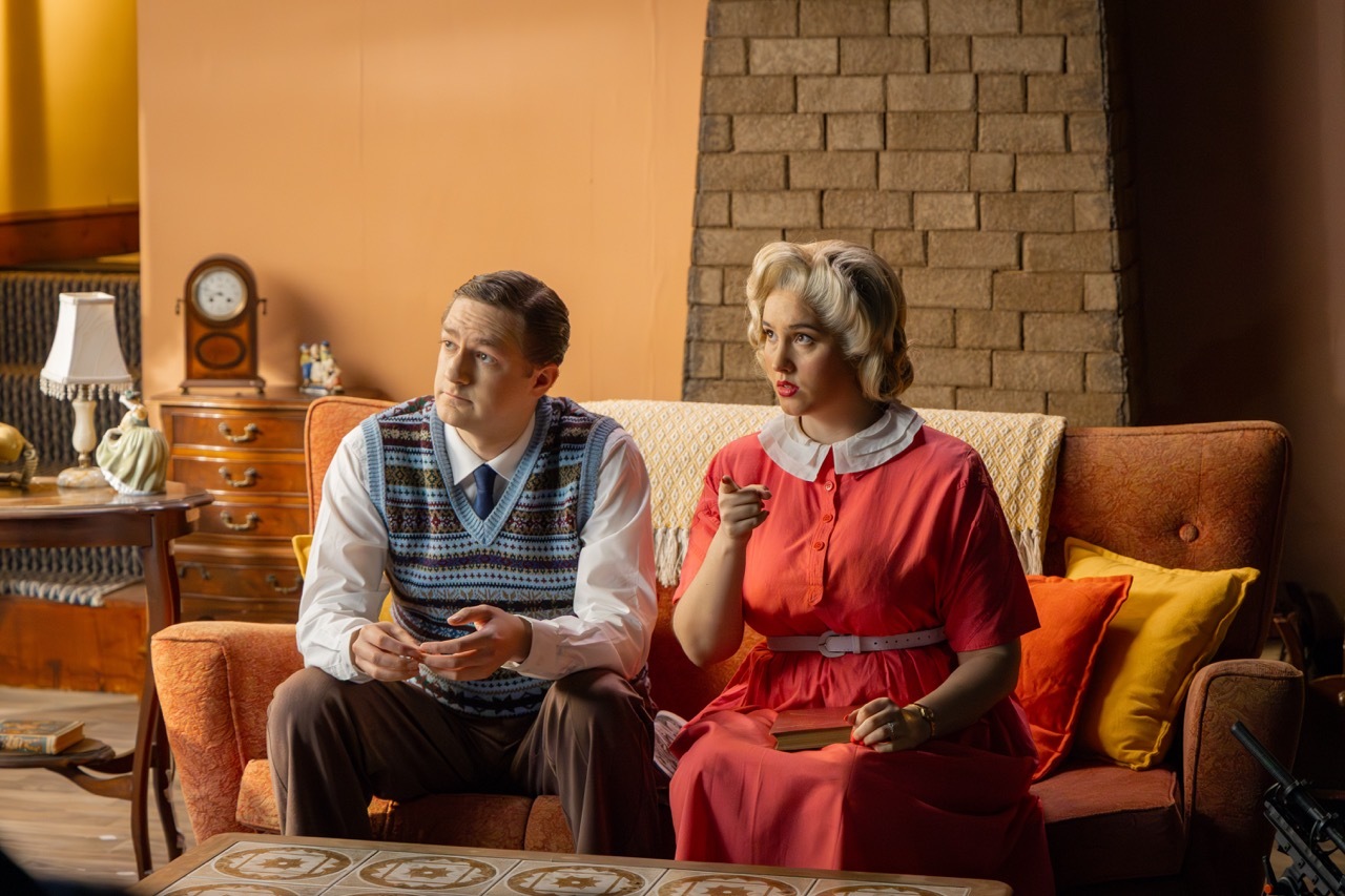 Elderly couple in a cosy living room, man in a sleeveless jumper and woman in a red dress, sitting on a sofa surrounded by wooden furniture and a brick fireplace.