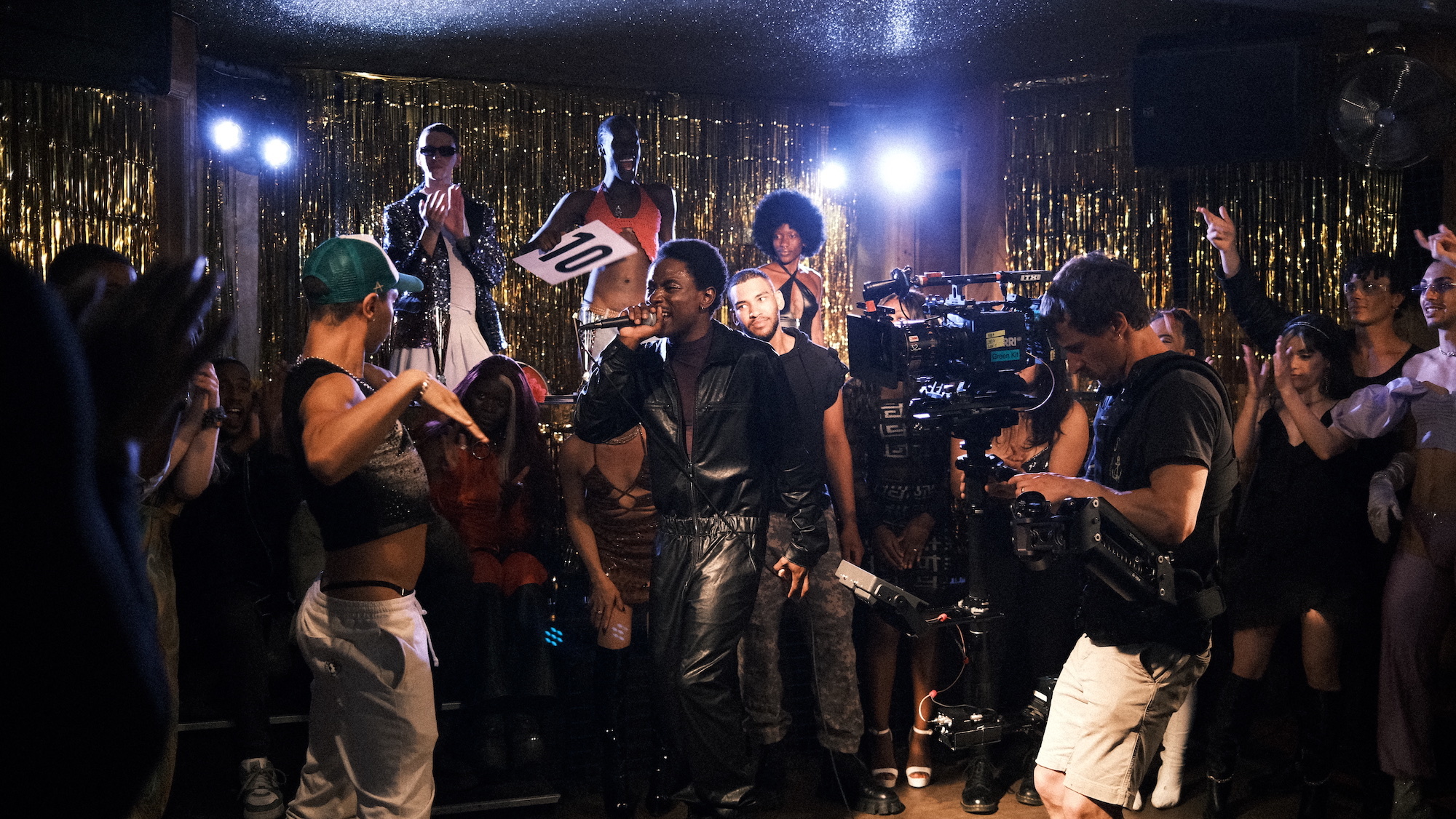 Scene from the film "Realness with a Twist" being filmed in a nightclub. A man looks through the viewer of a film camera surrounded by actors.