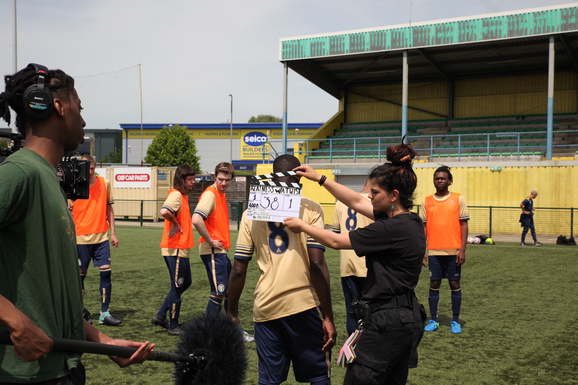 On a football field, a member of the crew for Realness with a Twist holds a clapper board behind an actor dressed in a football kit. Another crew member in the foreground holds a boom mic.
