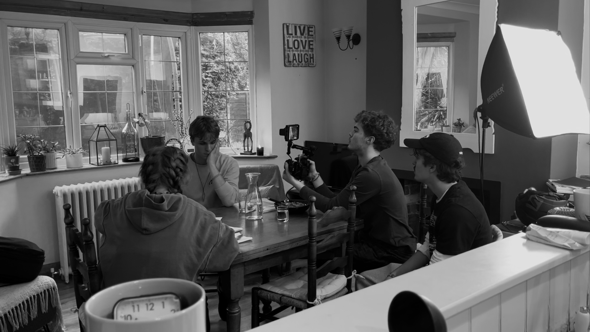 Black and white image of people seated around a dining table in a bright room with large windows, filming equipment visible.