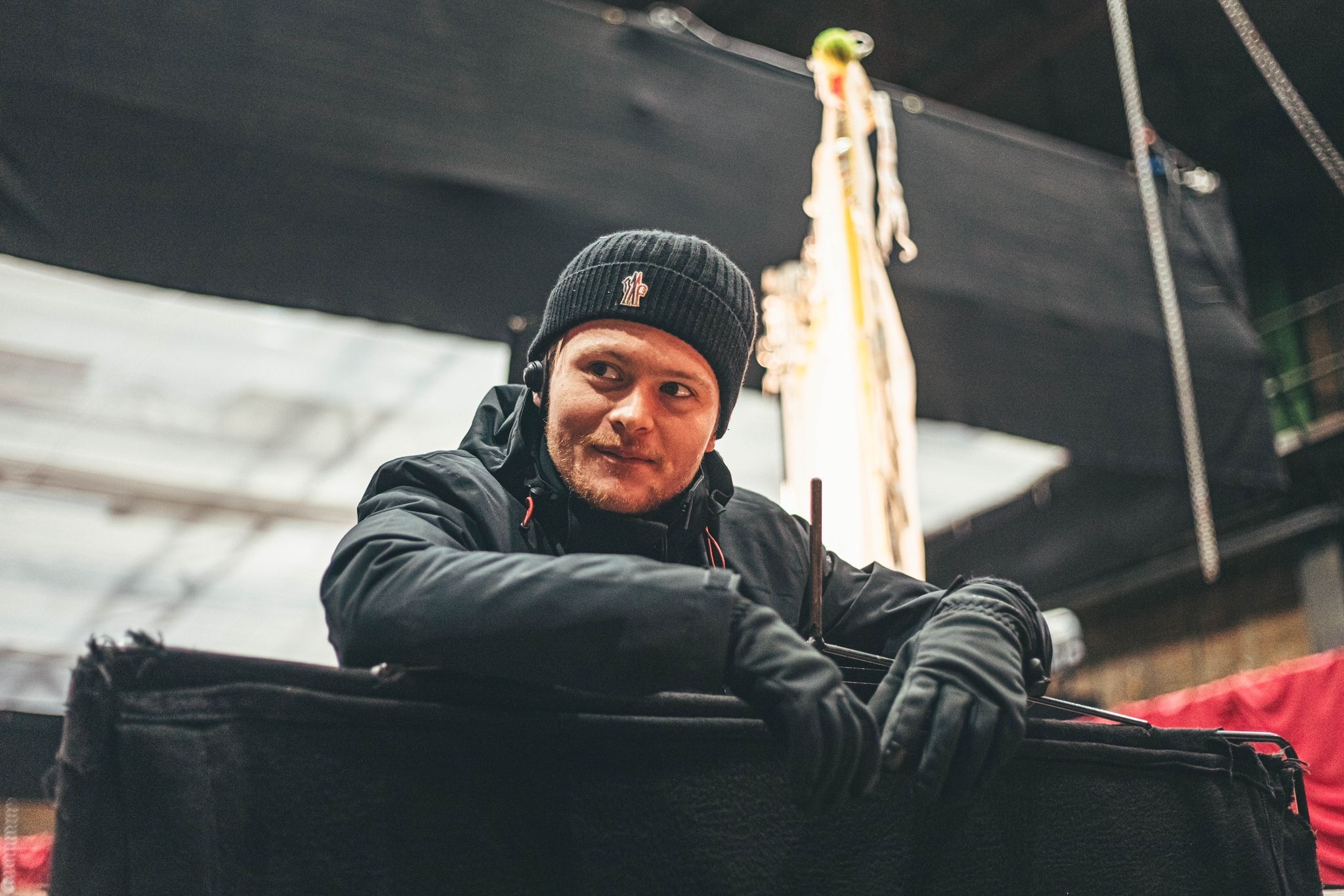 Man in dark winter clothing and beanie smiling whilst leaning on boat railing, with mast and rigging visible behind him.