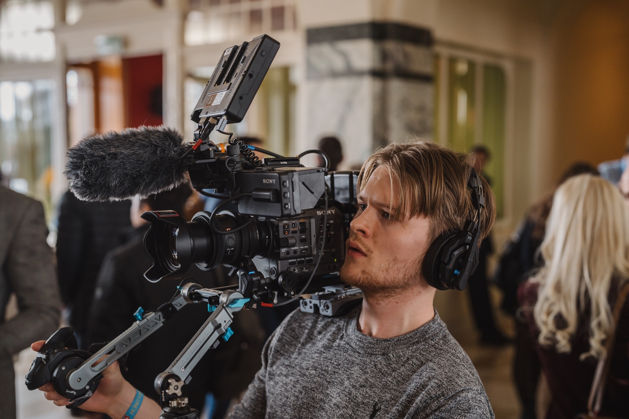 J Rigby operating professional film camera with boom microphone attachment in indoor setting with blurred people in background.