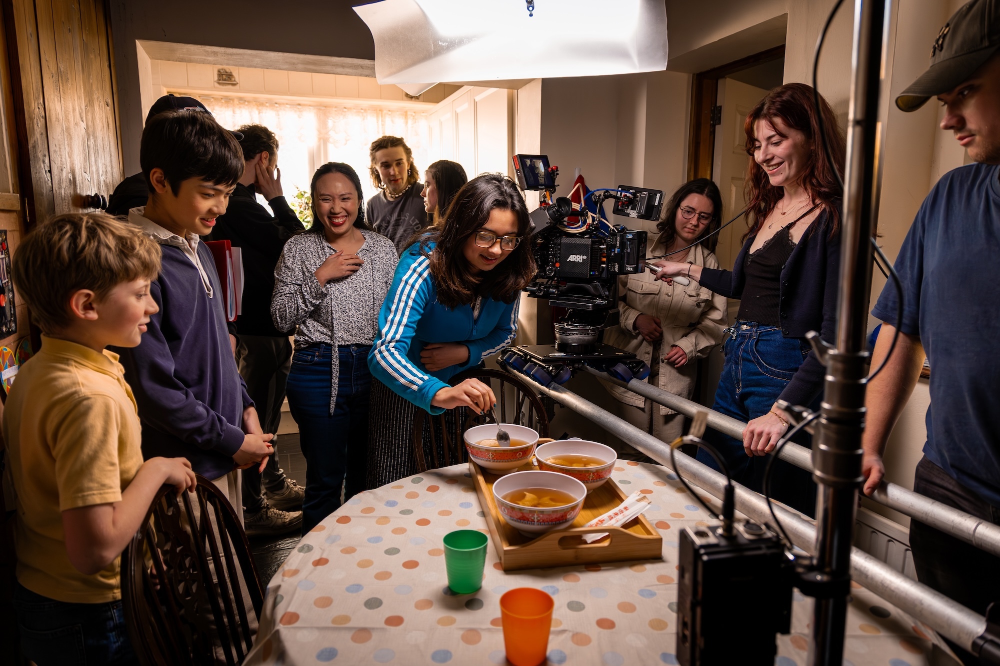 Group of BA (Hons) Film Production students and actors gathered around kitchen table with film equipment, woman in blue tracksuit preparing food whilst being filmed.
