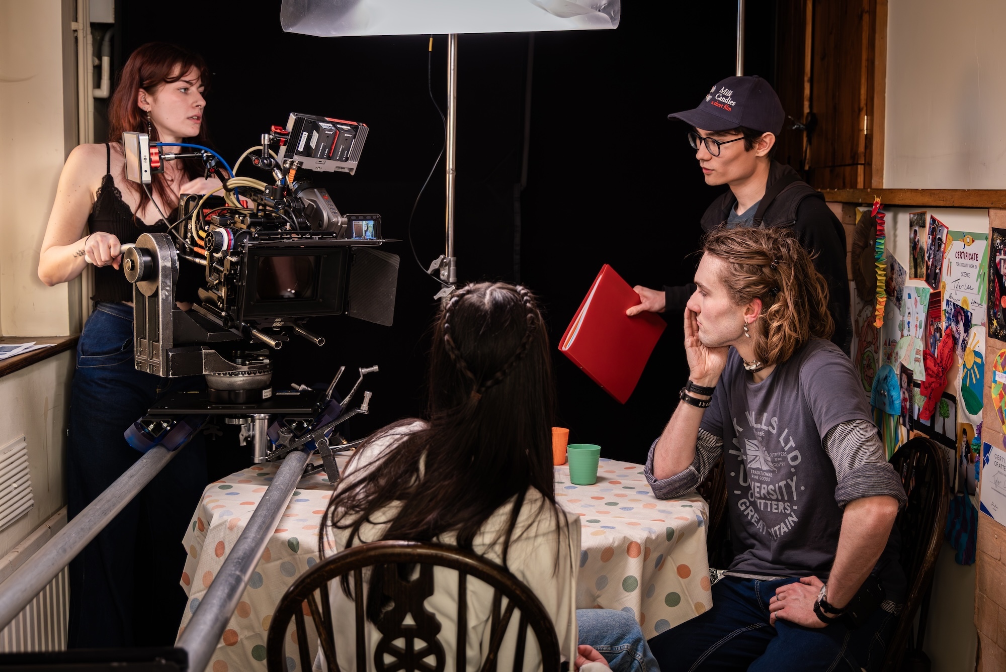 Crew of BA (Hons) Film Production students in domestic setting: person operating camera on left, person with cap holding red folder centre, person with long hair seated right.