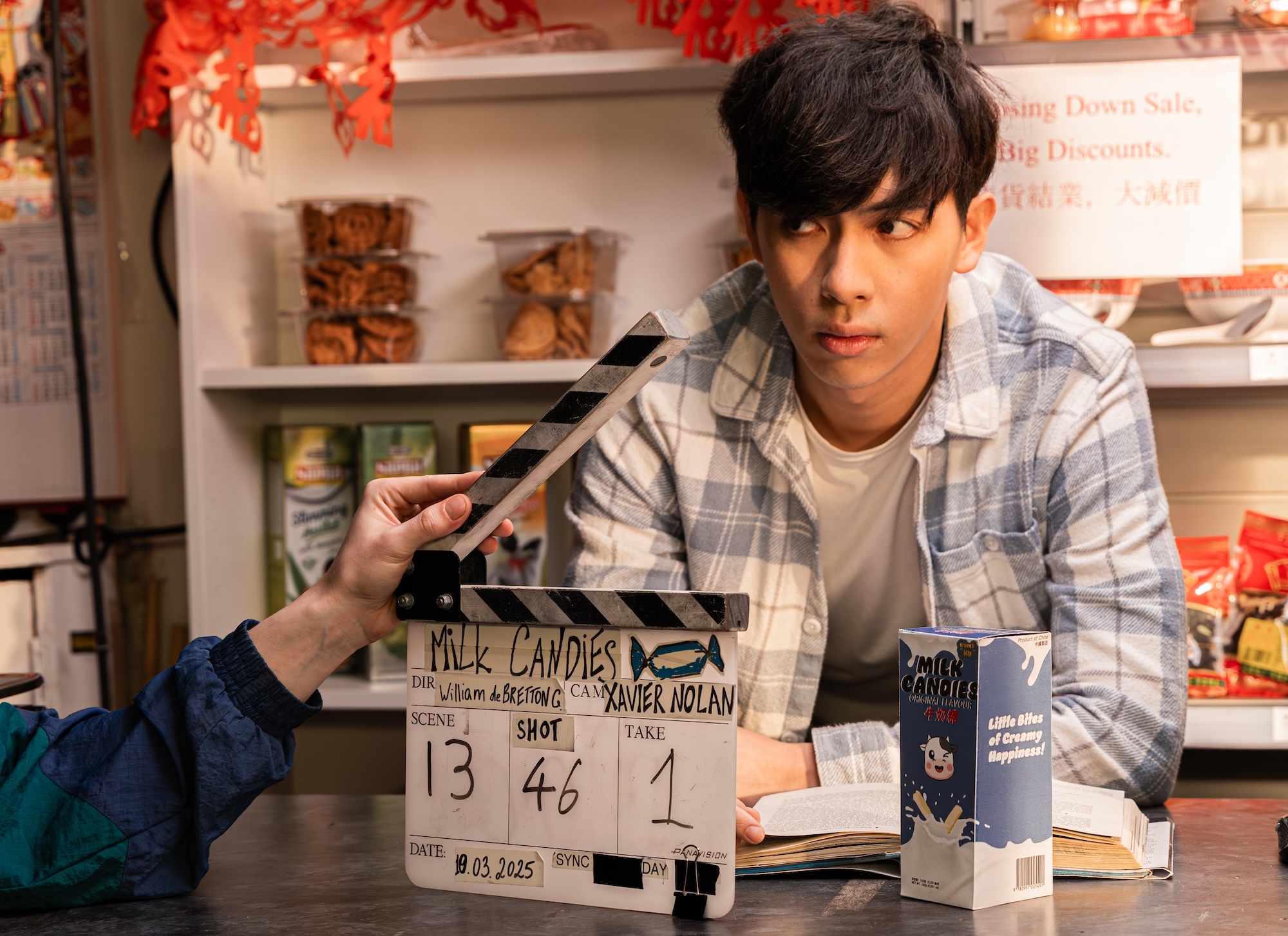 Actor L Hong sits at table with film clapperboard showing scene 13, take 46. Shelves with food items visible behind.