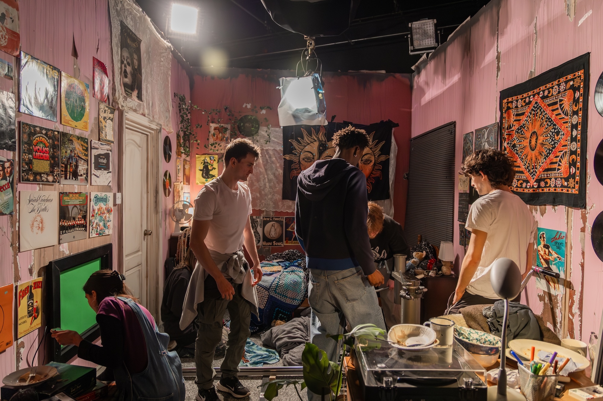 Pink-walled bedroom with artwork covering walls, three people standing, one sitting on floor, bed with colourful bedding, cluttered surfaces.