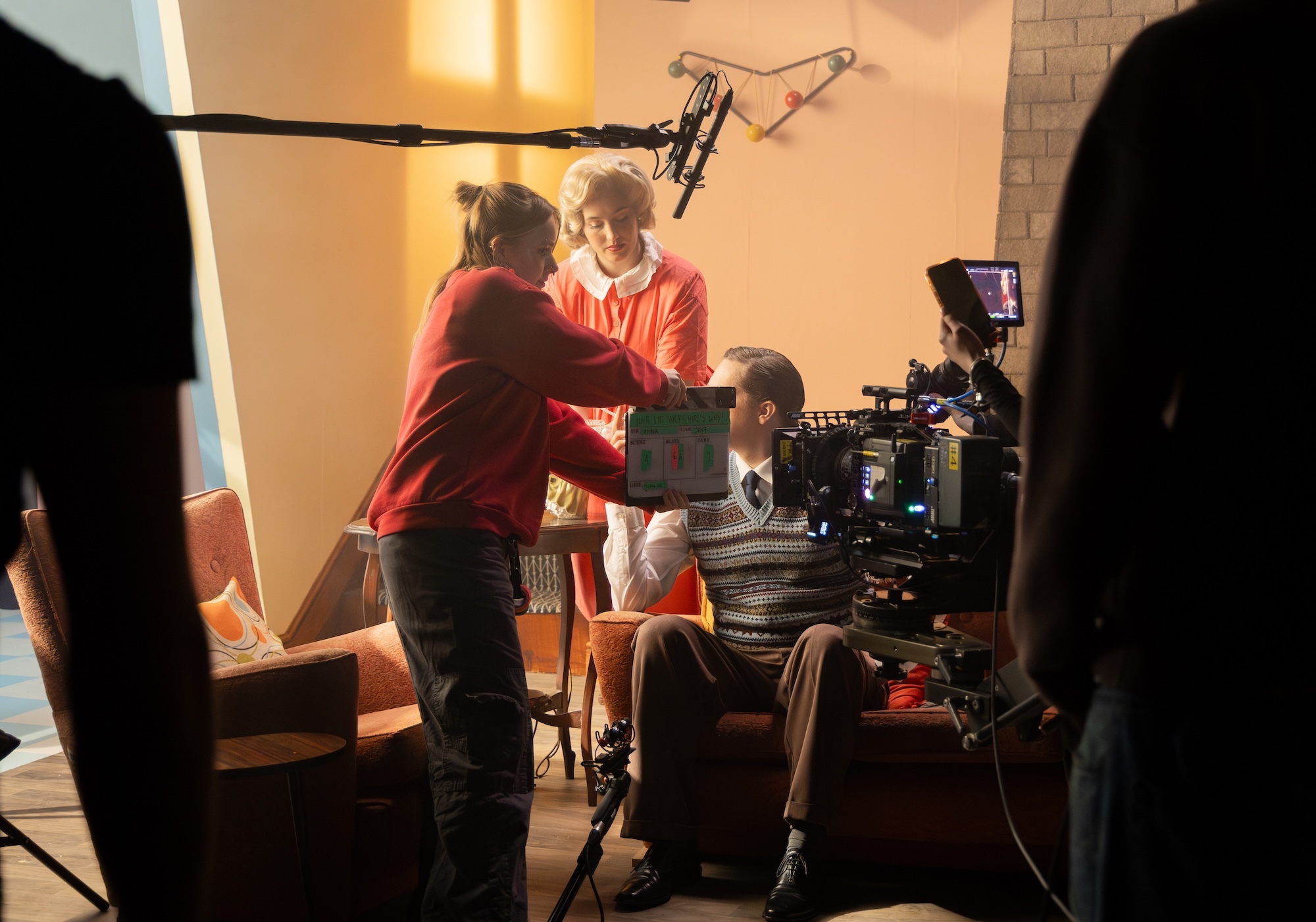 Film set with crew member adjusting actor in red jumper, camera equipment, boom mic, and warm lighting in brick-walled room.