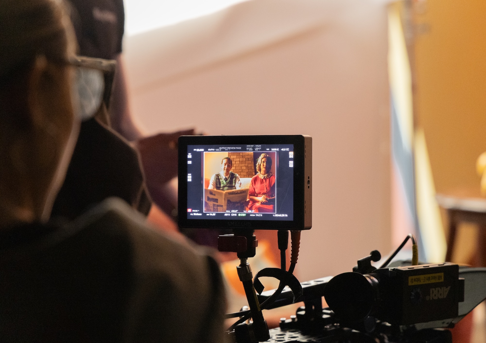 Camera operator filming with professional video equipment, small monitor displaying two people in orange/red setting visible on rig.