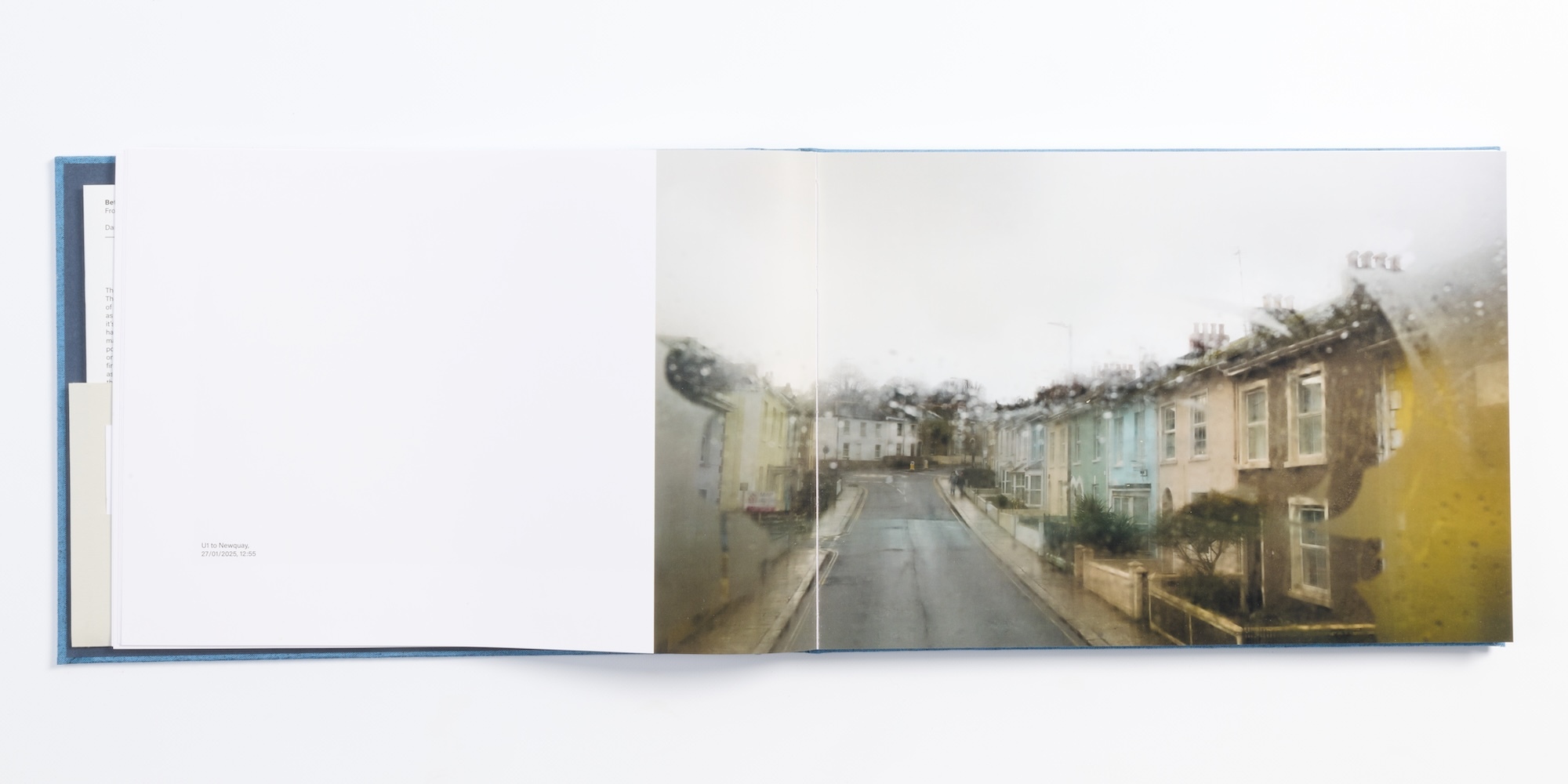 Open book with blank white left page and right page showing watercolour street scene in muted greys, blues, and yellow tones.