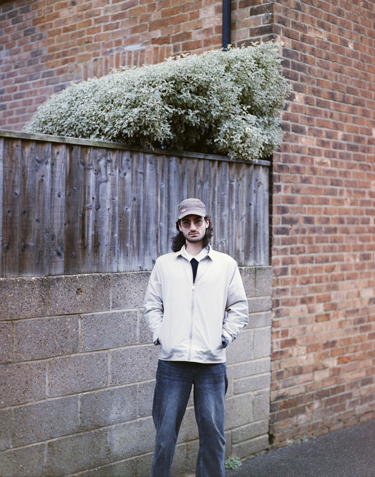 Portrait of G Buca standing with hands in pockets in front of a brick wall with a small wooden fence above it.