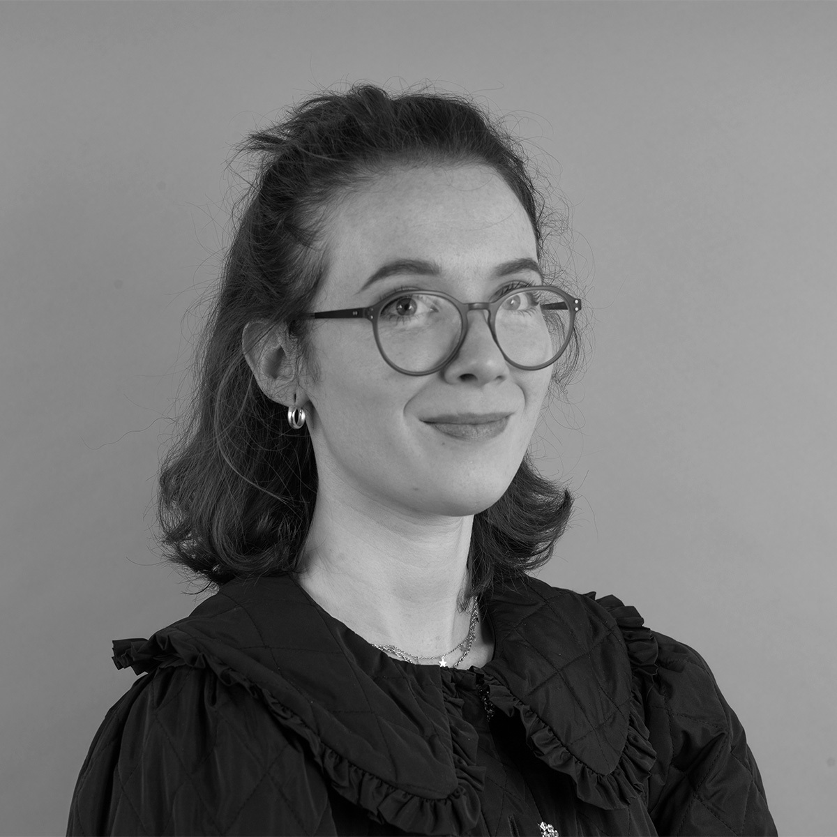 Black and white portrait of woman with shoulder-length hair, round glasses, and dark ruffled blouse against plain background.