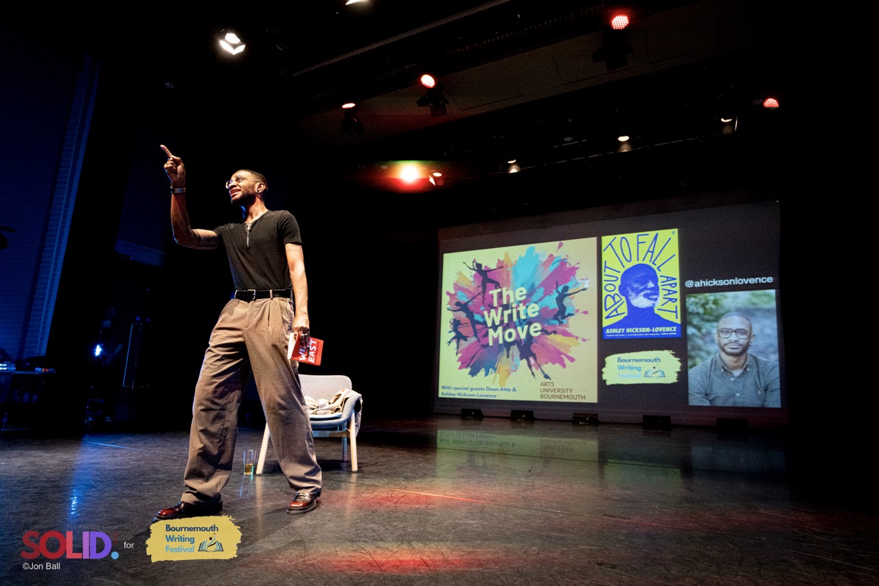 Ashley Hickson-Lovence stood on-stage wearing a black t-shirt and brown trousers, pointing to the audience. Lights above him and 'The Write Move' poster projected in the background, colourful explosion of dance on yellow.