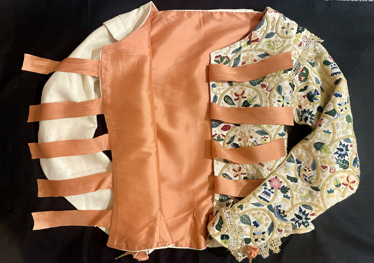 A 1610s jacket resting on a plain background unfastened and open showing the inner lining.
