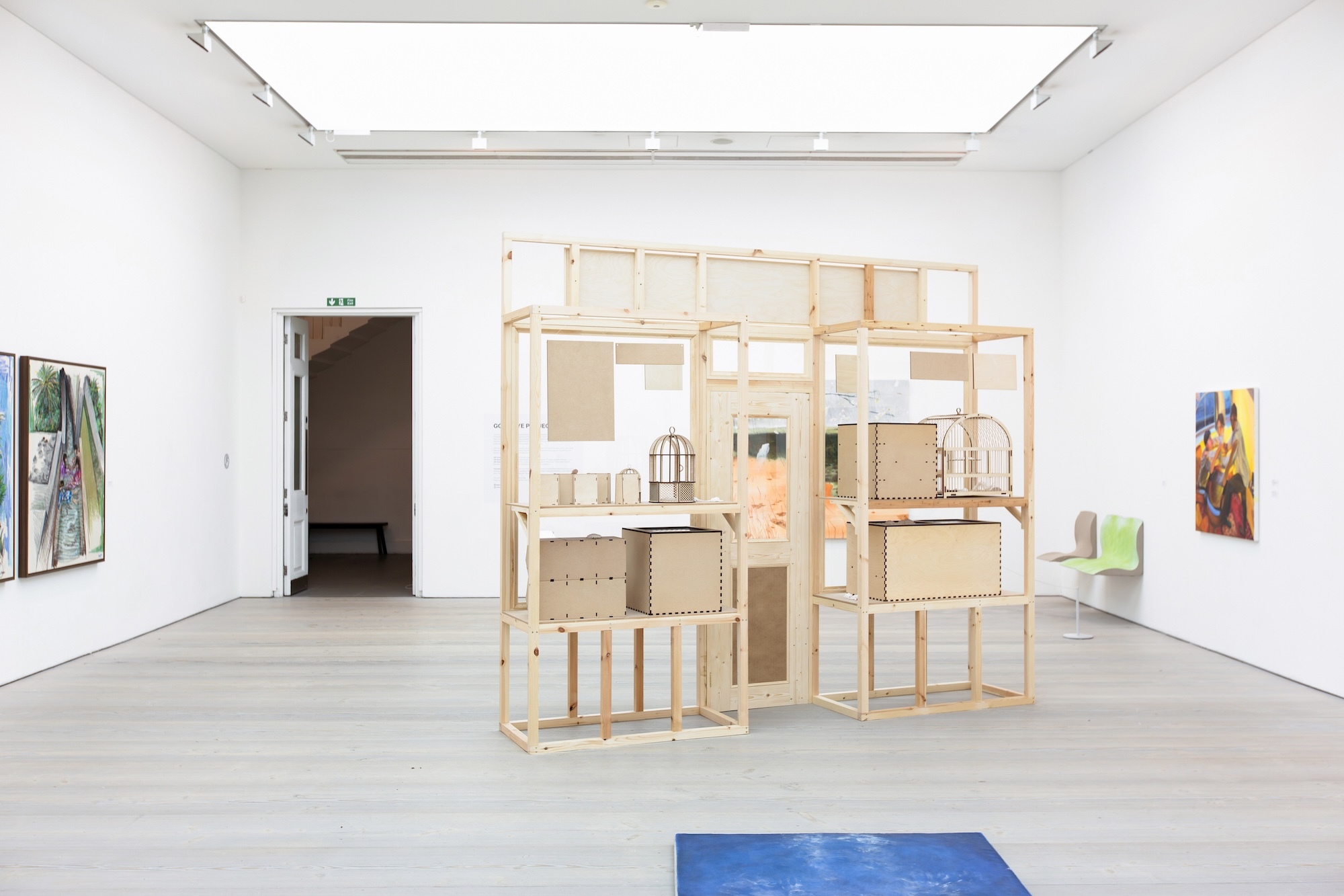 White gallery space with wooden shelving structure displaying various objects in centre, paintings on walls, skylight above.