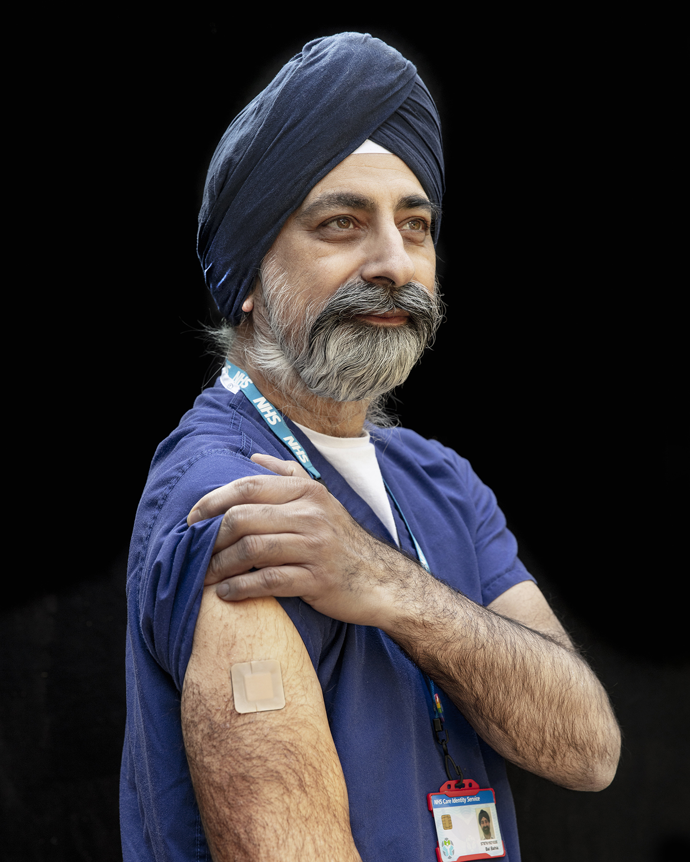 A man with a turban looks off to the right and lifts their sleeve up to show a small plaster on their arm.
