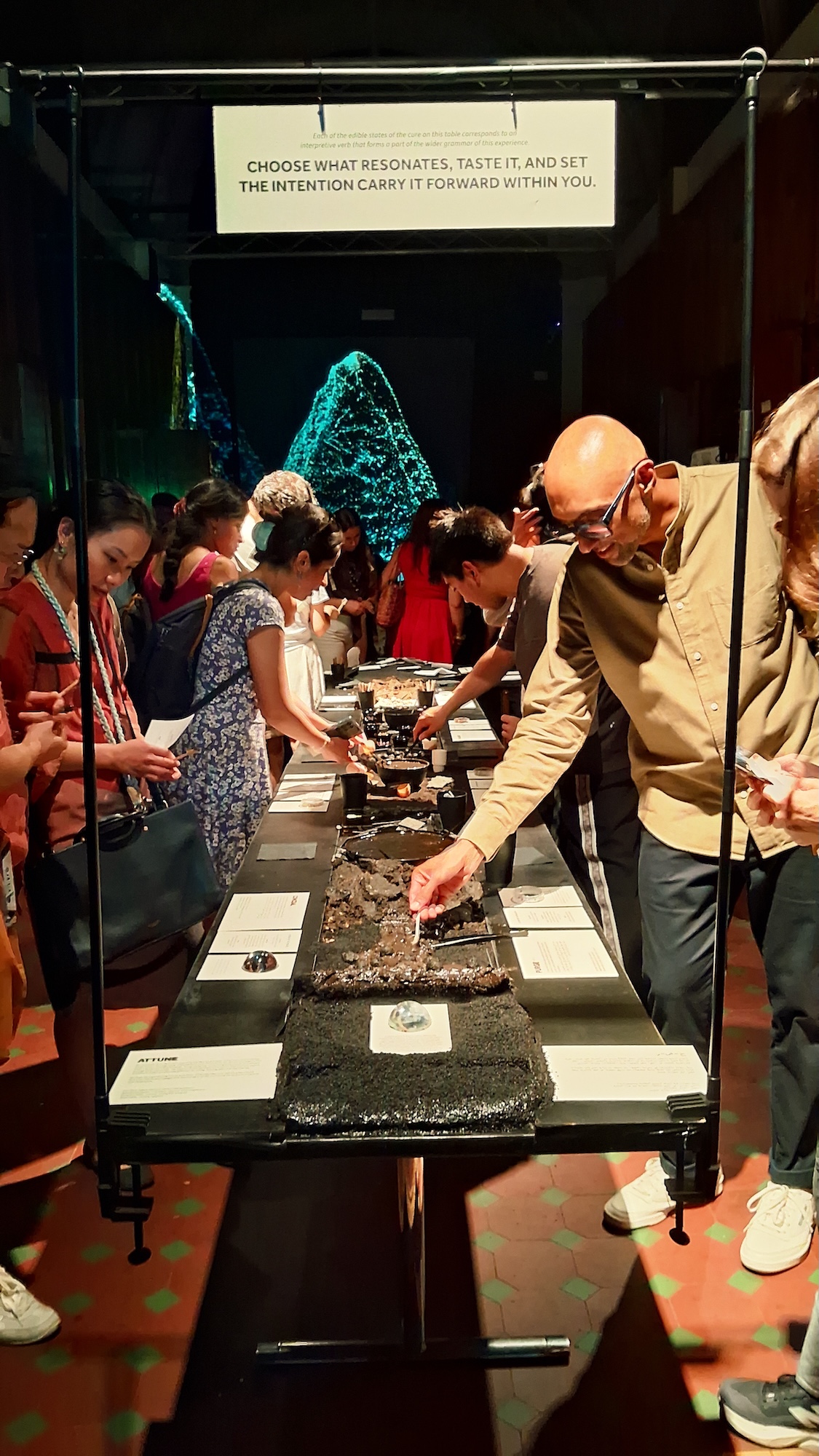 A man in a beige jacket leans over a long tasting table while several people sample food at an indoor event, with a sign above and a glowing cone-shaped display in the background.