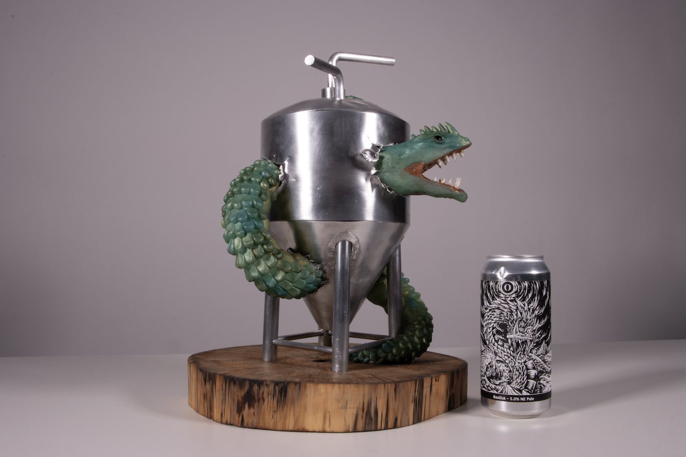 Model of a basilisk tearing through a metal tank on a wooden stand, with a beer can next to it.
