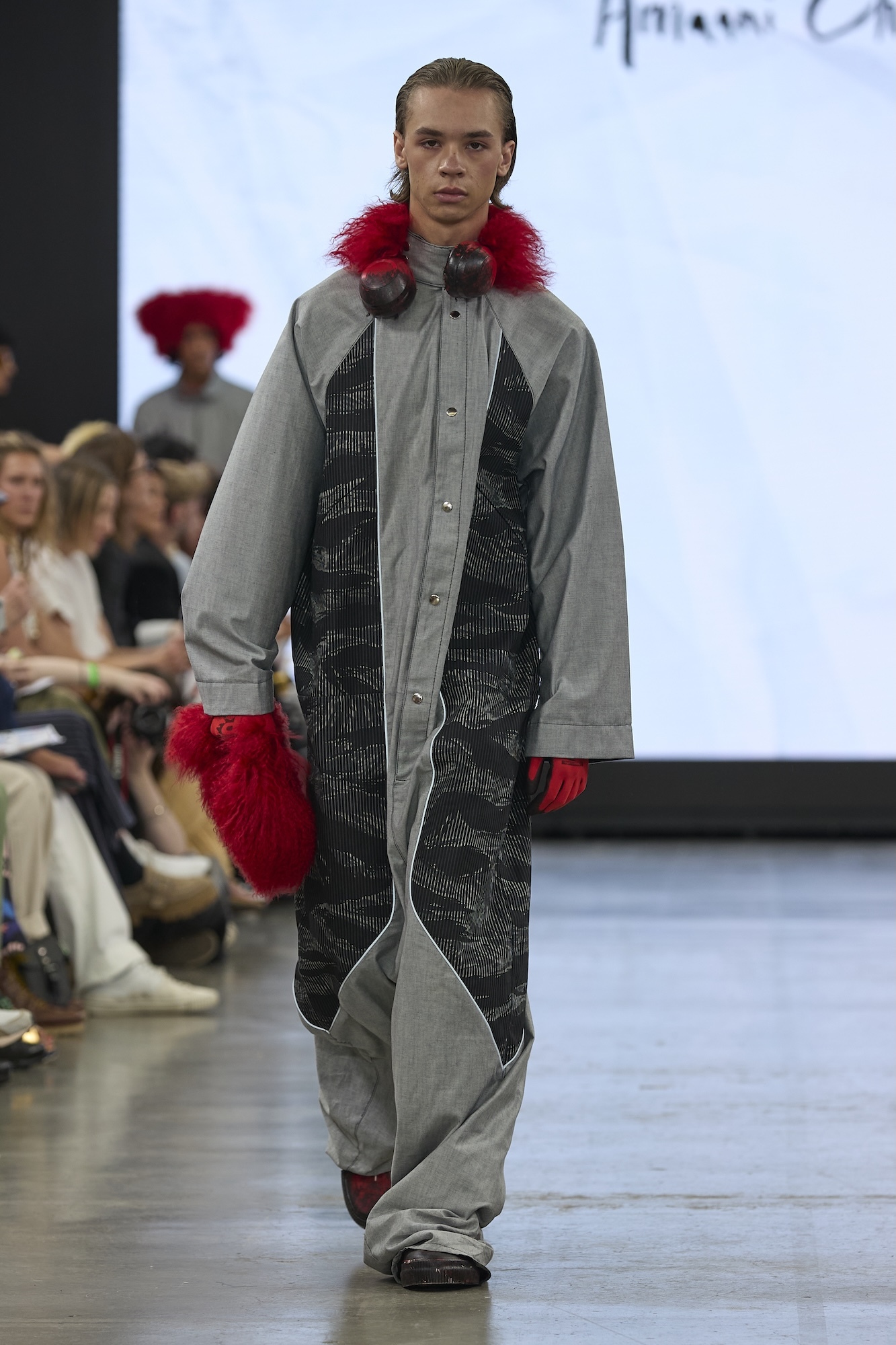 Male model on runway wearing grey tracksuit with red fur hood trim and black geometric panel insert, audience seated behind.