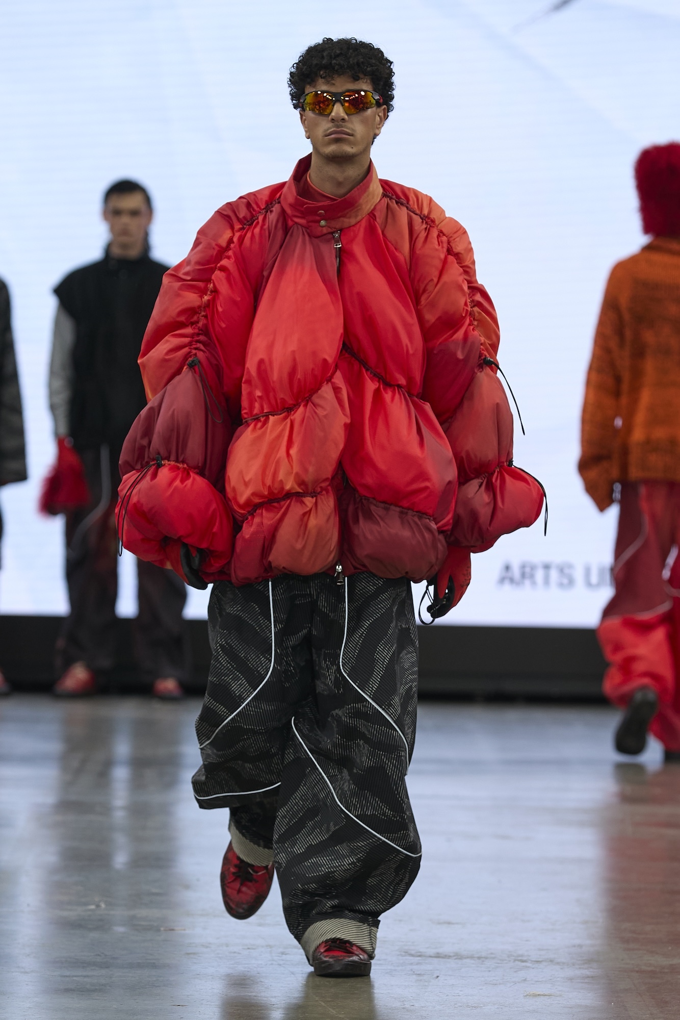 Male model wearing oversized red puffer jacket, dark trousers and red-tinted sunglasses walks down fashion runway.