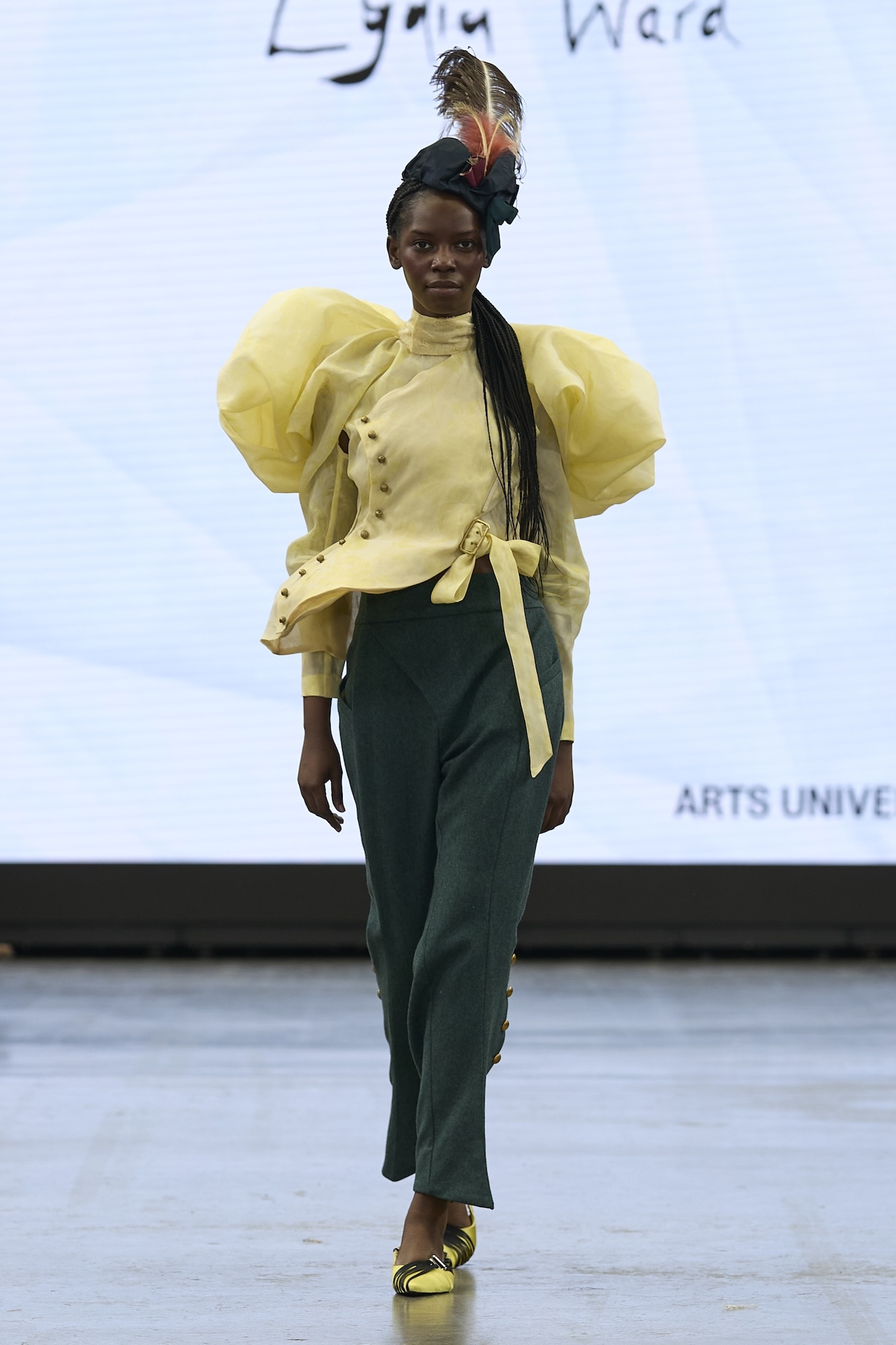 Black woman on catwalk wearing yellow puffy-sleeved blouse, dark green trousers, feathered headpiece, and yellow strappy shoes.