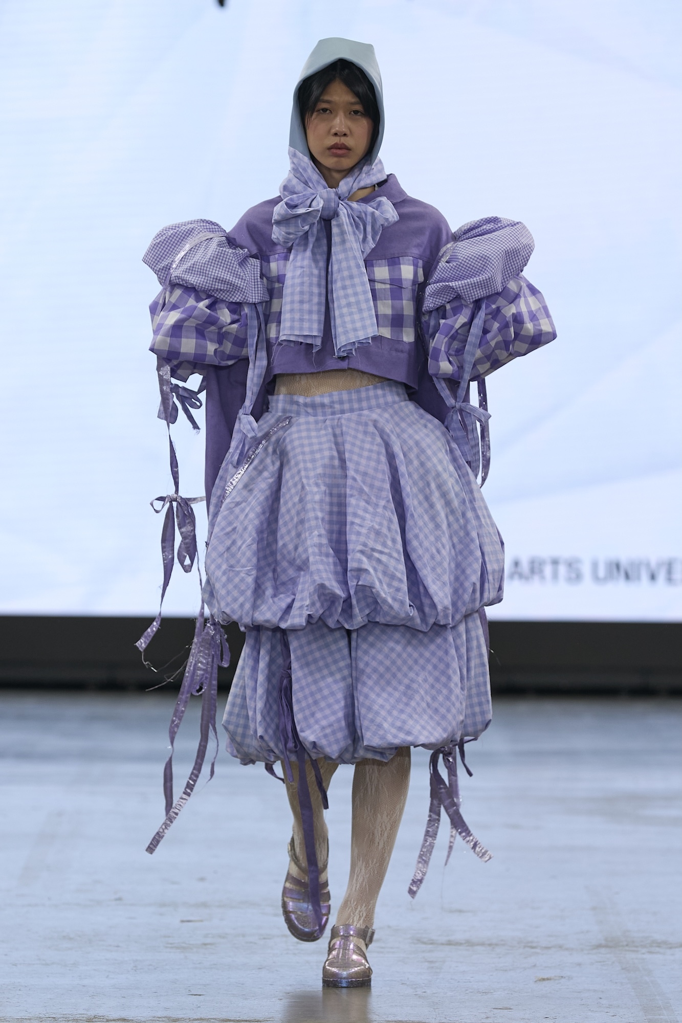 Model on catwalk wearing layered purple and white checkered outfit with oversized puffy sleeves, headscarf, and platform shoes.