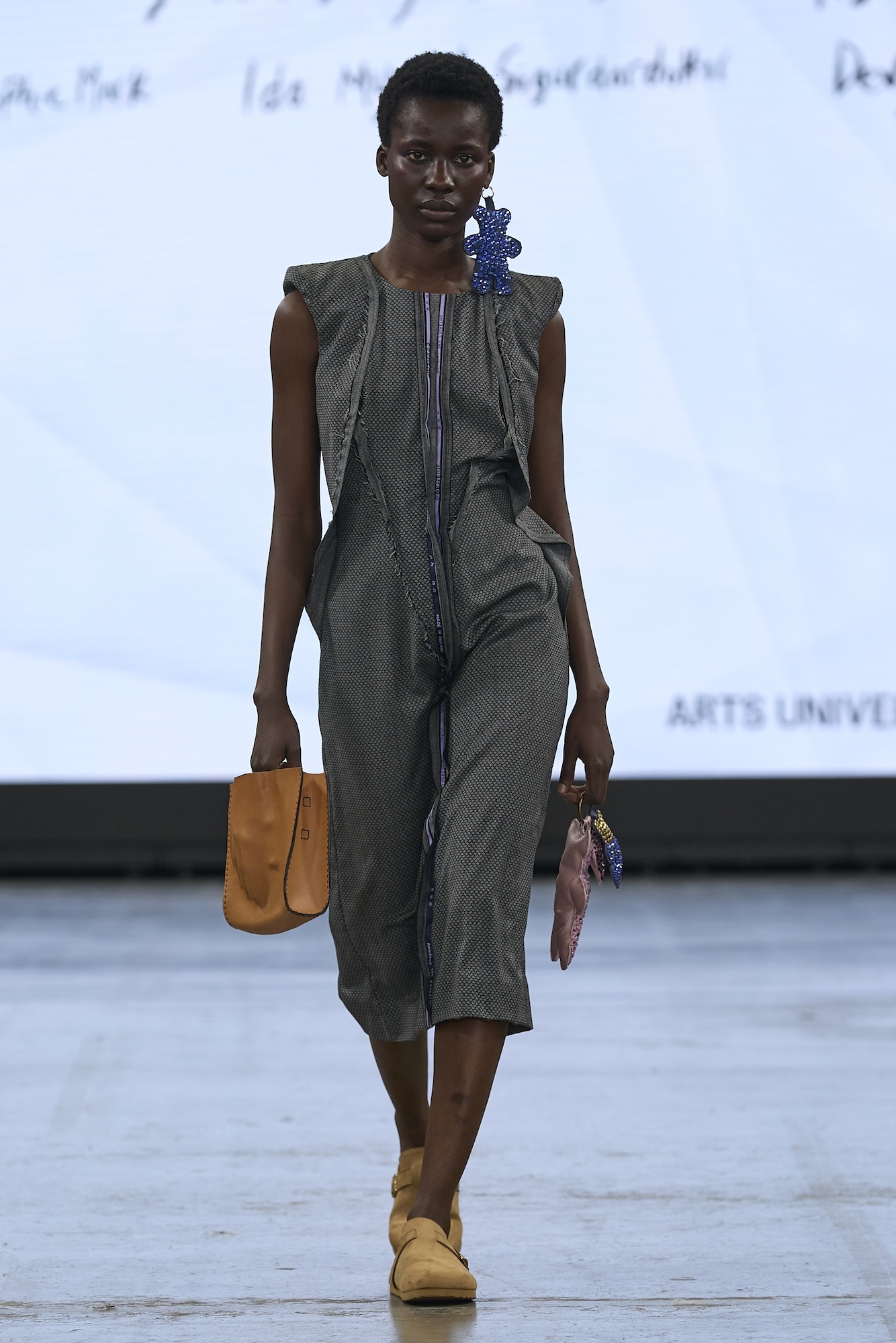 Model on runway wearing grey sleeveless jumpsuit with front zip, blue beaded earring, carrying tan leather bag and brown clutch.