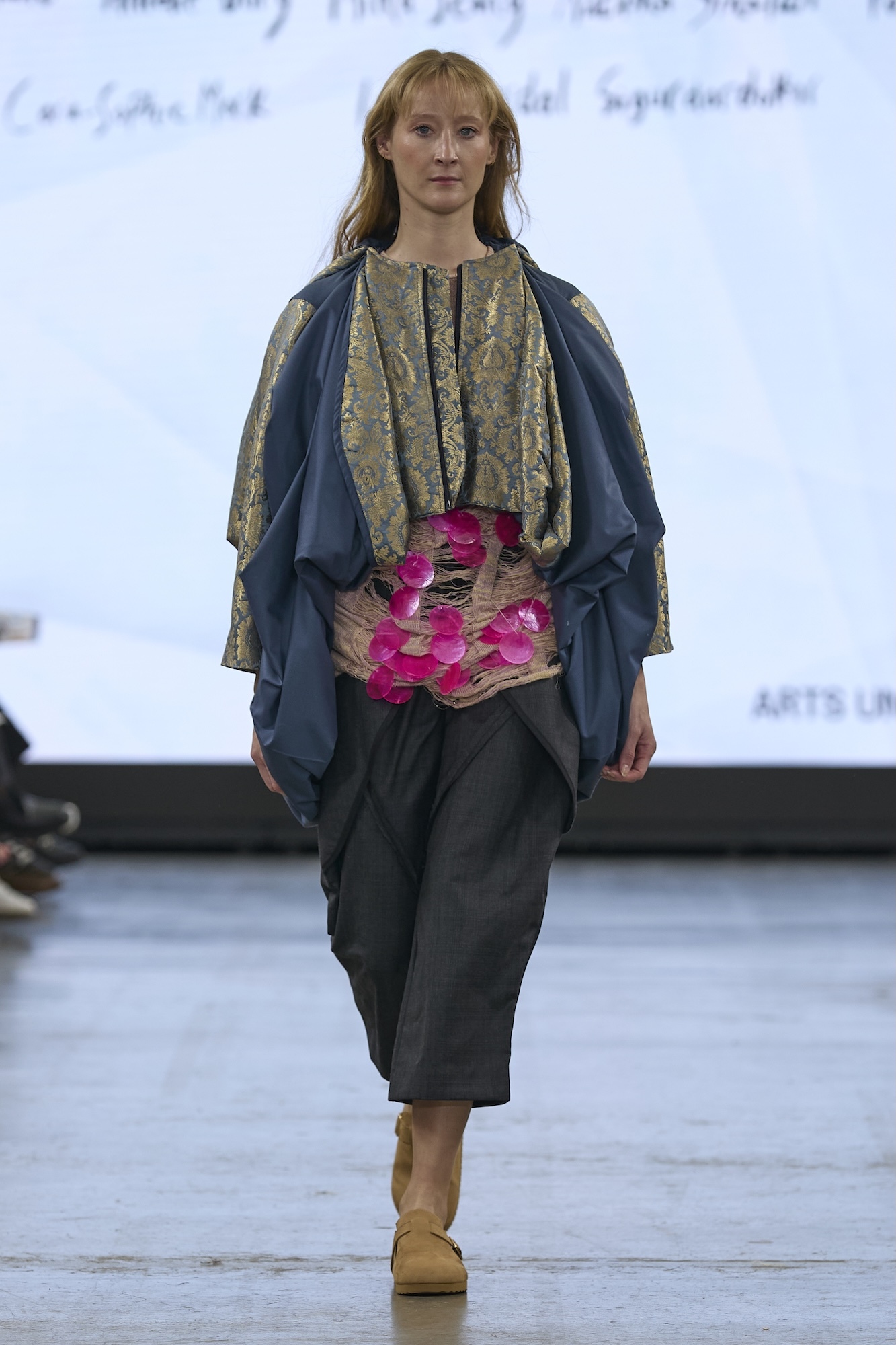 Model on runway wearing layered outfit: blue cape over patterned top, woven basket with pink flowers at waist, dark green skirt.