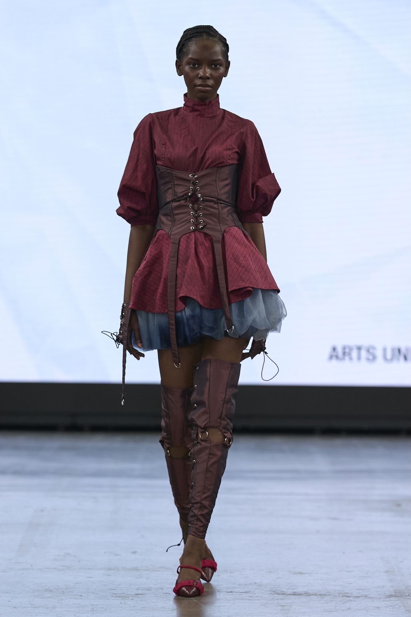 Model on runway wearing burgundy blouse with brown leather corset, blue tulle skirt, and tall lace-up boots against white backdrop.
