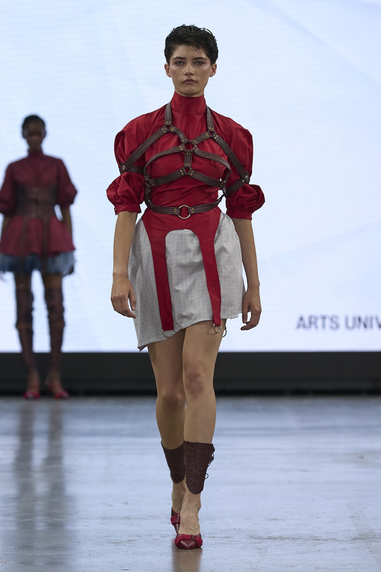 Model on runway wearing red top with brown leather harness straps, white skirt with red panels, and brown leather leg warmers.