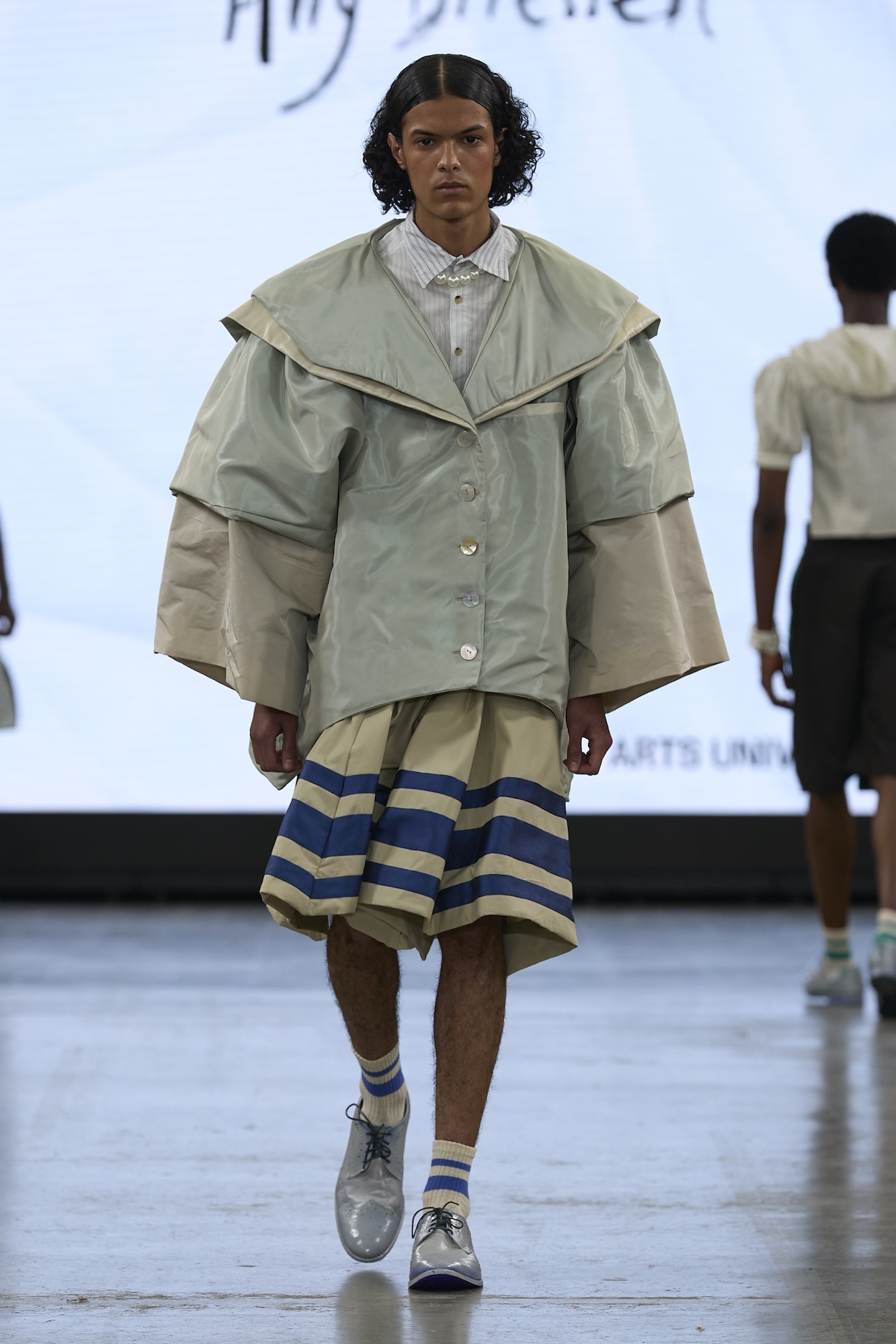 Model on runway wearing oversized sage green jacket over white shirt, yellow shorts with blue stripes, and grey trainers.