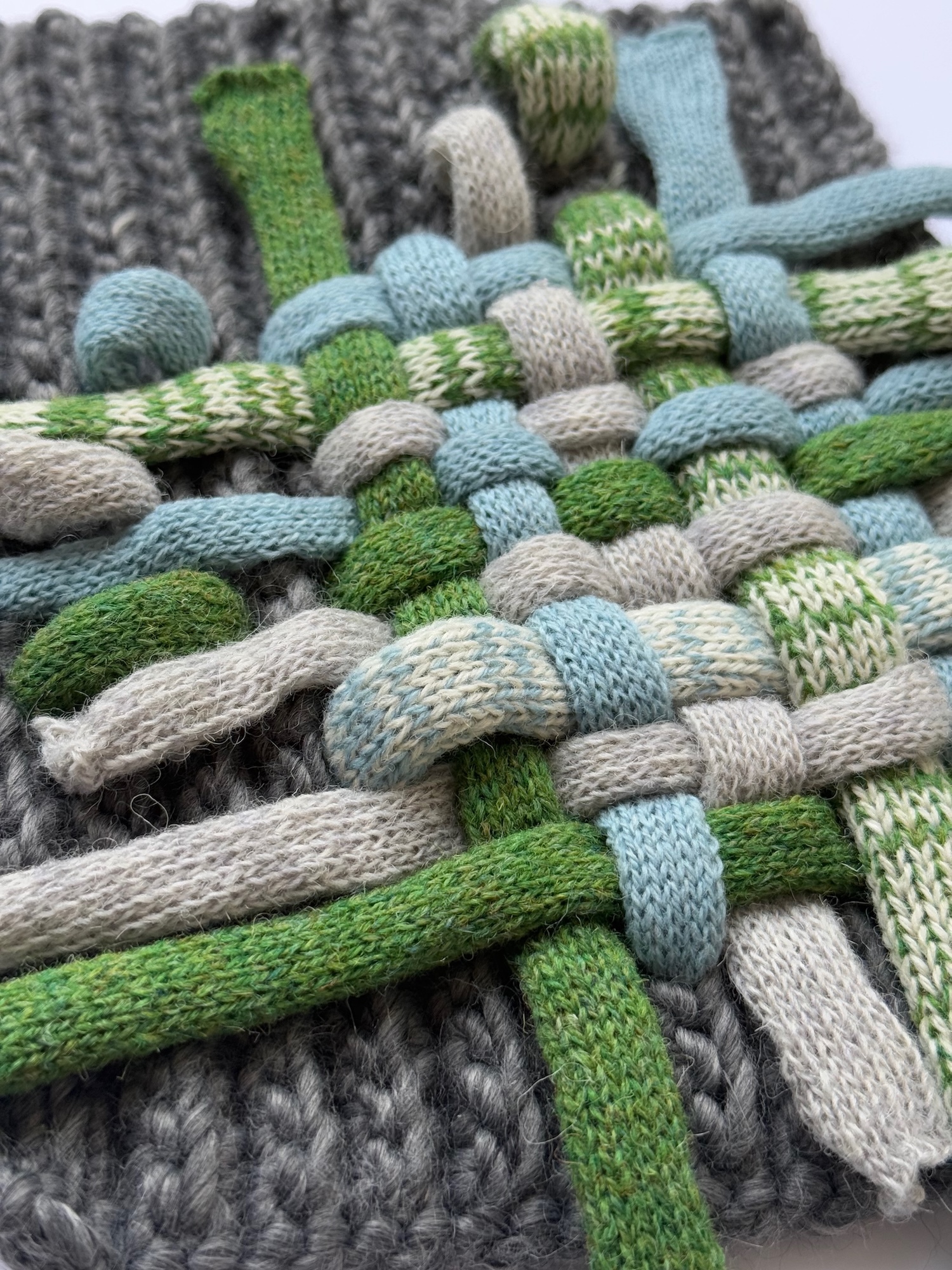 Knitted strips in green, blue, beige and grey woven together in basket-weave pattern on grey knitted background.